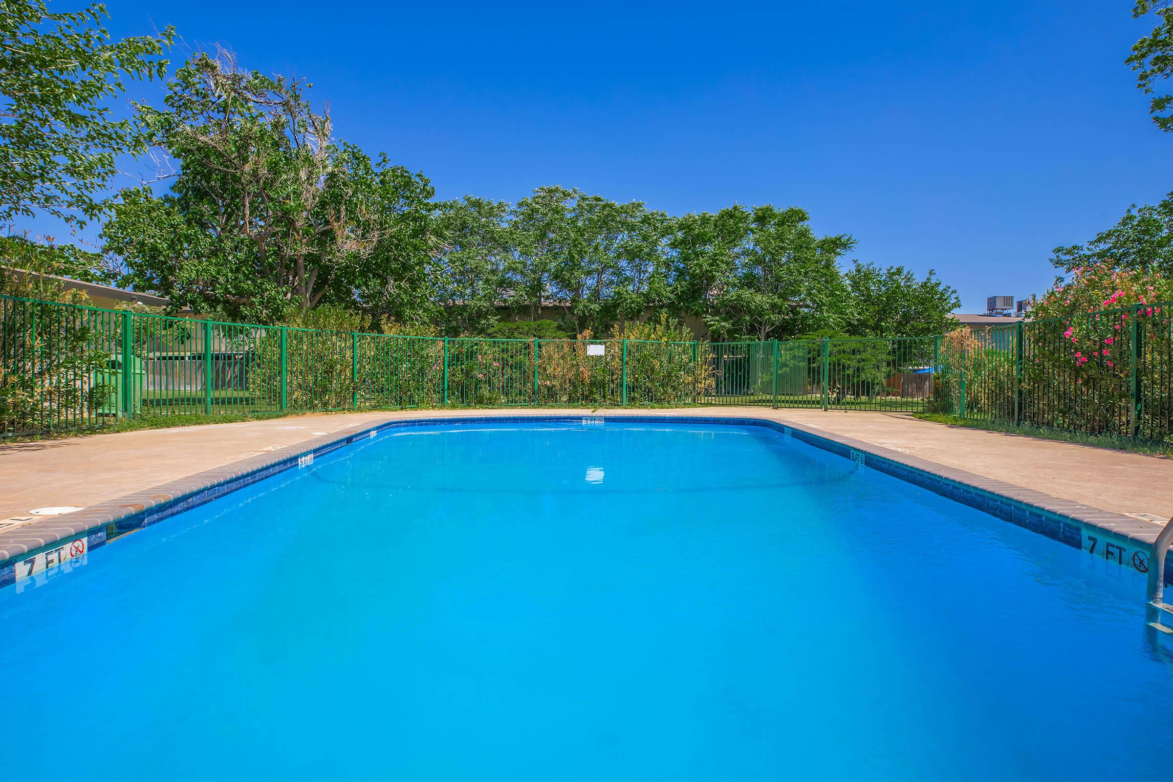 A clean, inviting swimming pool surrounded by a green fence and lush trees. The clear blue water reflects the bright sky, indicating a sunny day. The area is well-maintained, creating a relaxing atmosphere for outdoor enjoyment.