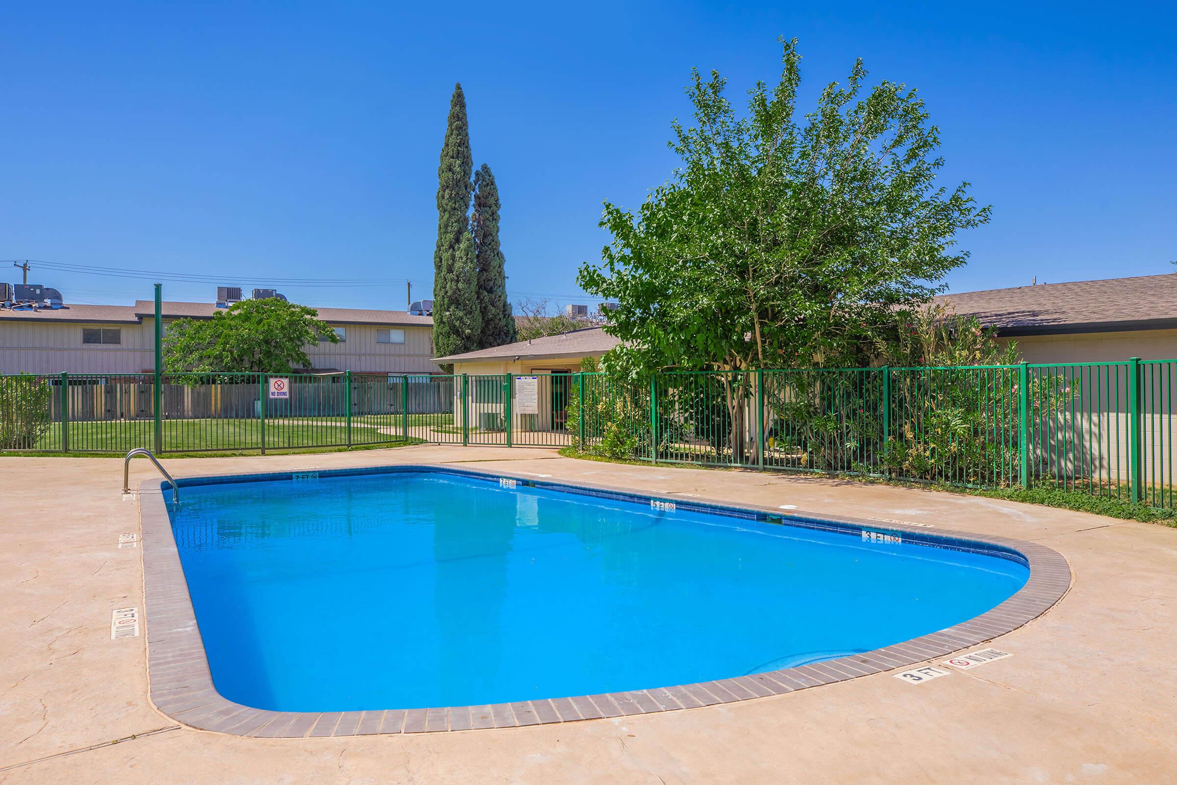A bright blue swimming pool surrounded by a paved area and green fences. Lush trees and shrubs are visible nearby, with a clear blue sky above. The area appears well-maintained and inviting, suitable for relaxation or recreation.