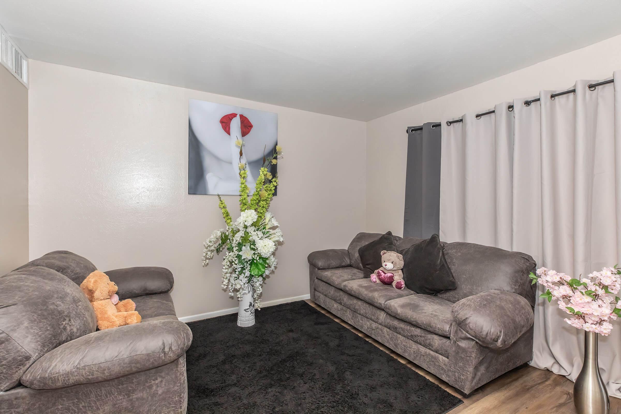 A cozy living room featuring two gray sofas with plush cushions. One sofa has a teddy bear sitting on it. A large floral arrangement is placed in a white vase, adding a touch of elegance. The walls are adorned with a large artwork, and curtains in soft tones frame a window, enhancing the warm atmosphere.
