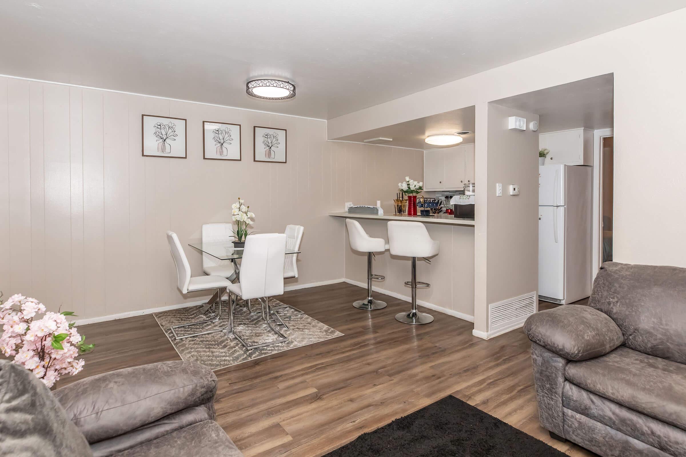 Modern living space featuring a dining area with a glass table and four white chairs, surrounded by light beige walls. In the background, a kitchen bar with three white stools and a refrigerator. A cozy lounge area with a gray sofa and a dark rug, complemented by decorative flower arrangements.