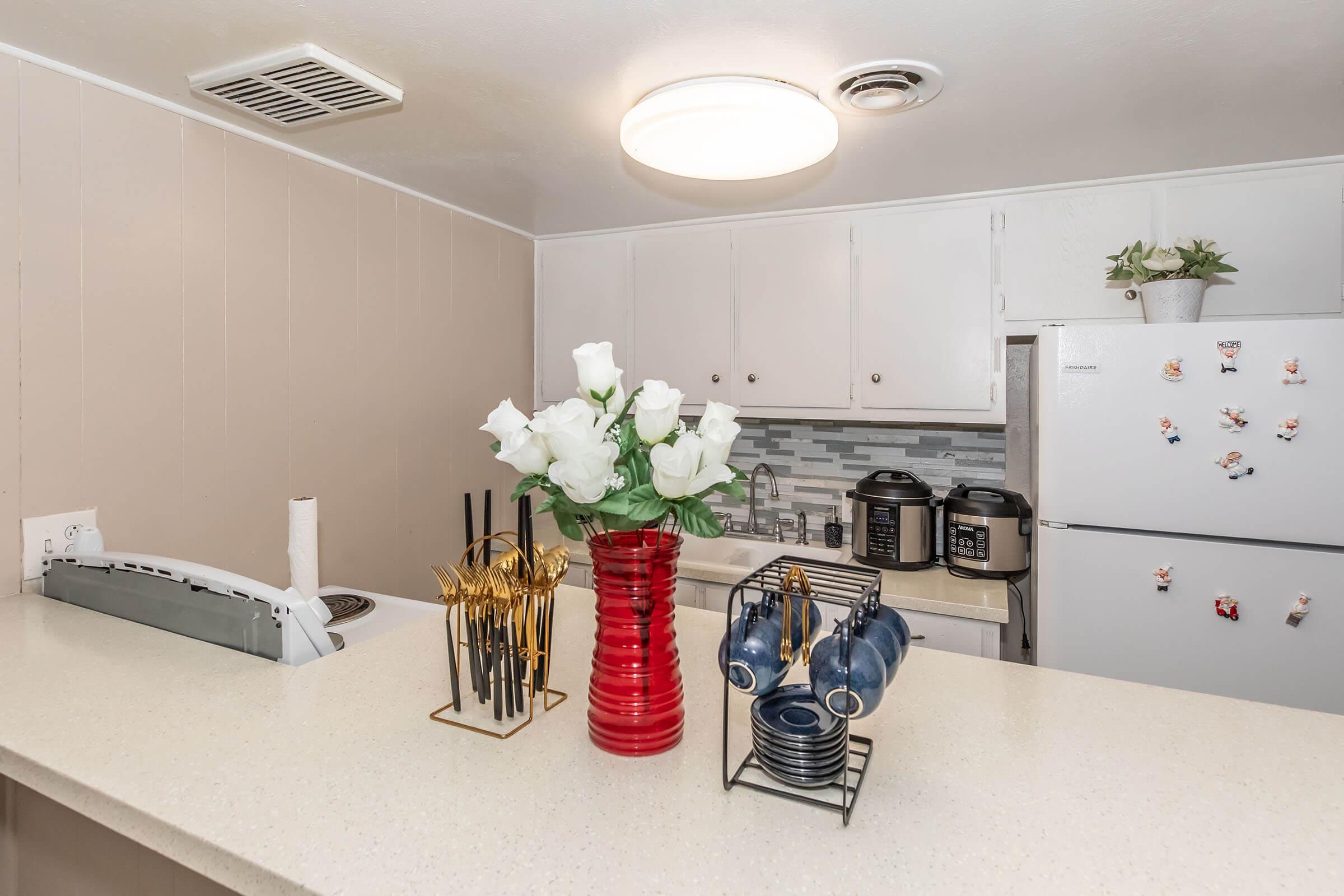 A modern kitchen counter featuring a red vase with white roses, a set of gold utensils in a black holder, and blue ceramic mugs on a rack. The background includes a white refrigerator and a tiled backsplash, creating a warm and inviting atmosphere.