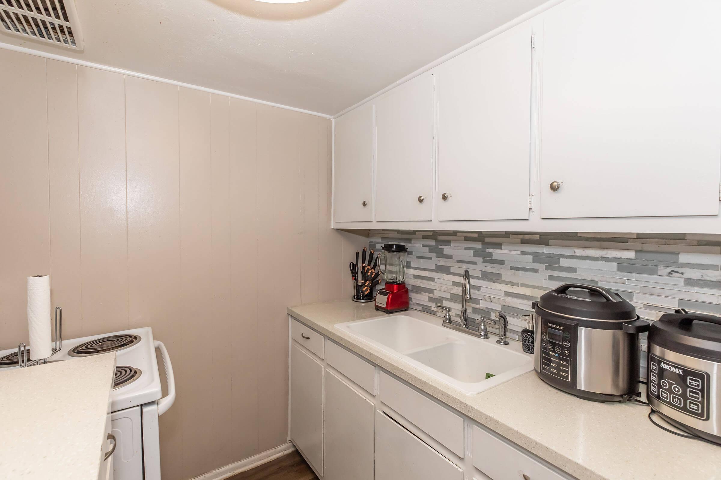 A compact kitchen featuring white cabinets, a light-colored countertop, and a modern backsplash with gray and white tiles. The kitchen includes an electric stove, a sink with a faucet, and two appliances: a pressure cooker and a multi-cooker. Natural light illuminates the space, enhancing its brightness.