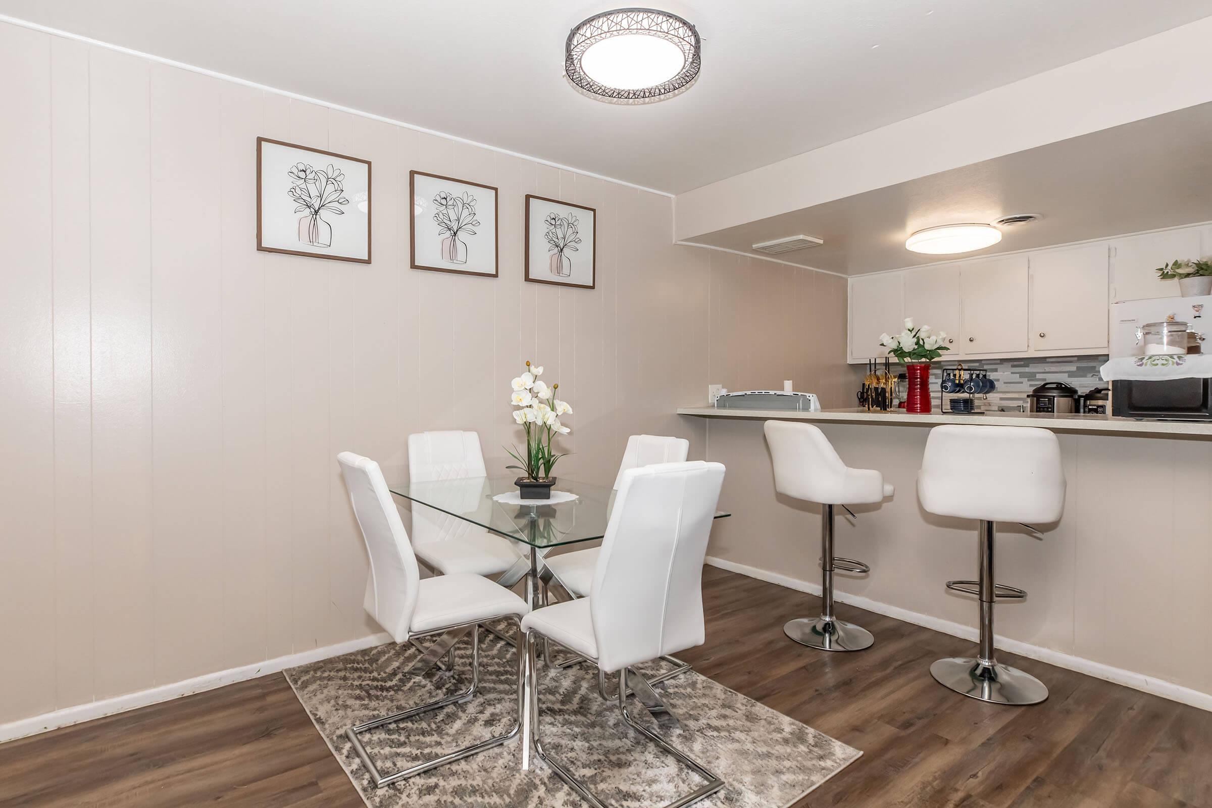 A modern dining area featuring a glass table with four white chairs, complemented by a decorative rug. In the background, there is a kitchen bar with high stools and decorative elements like flowers and framed artwork on the walls. Soft lighting adds a welcoming ambiance.