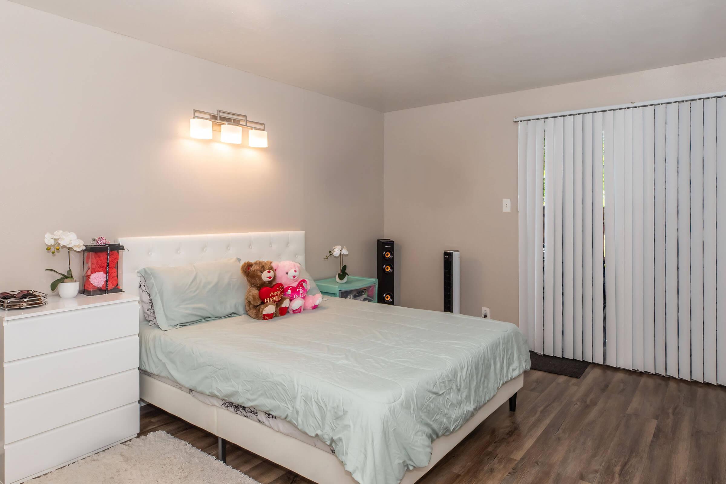 A cozy bedroom featuring a light blue bedspread with two teddy bears and a decorative pink bunny on the bed. There’s a white dresser beside the bed and a small pink table with a flower arrangement. A sliding door with vertical blinds leads to an outdoor area, and soft wooden flooring completes the look.