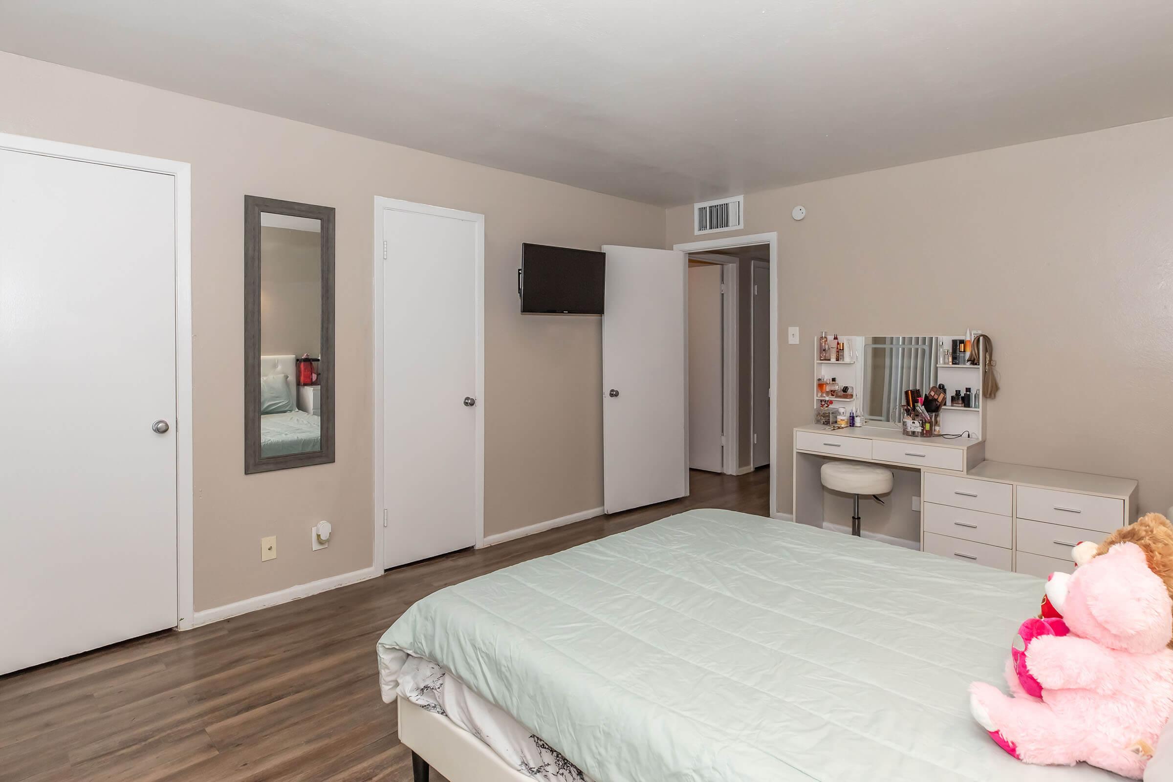 A cozy bedroom featuring a bed with a light teal comforter. There's a mirrored dresser with a vanity and a chair, and a wall-mounted TV. Two doors lead to adjacent rooms, and a pink stuffed animal is placed on the bed. The walls are painted a warm beige, creating a inviting atmosphere.