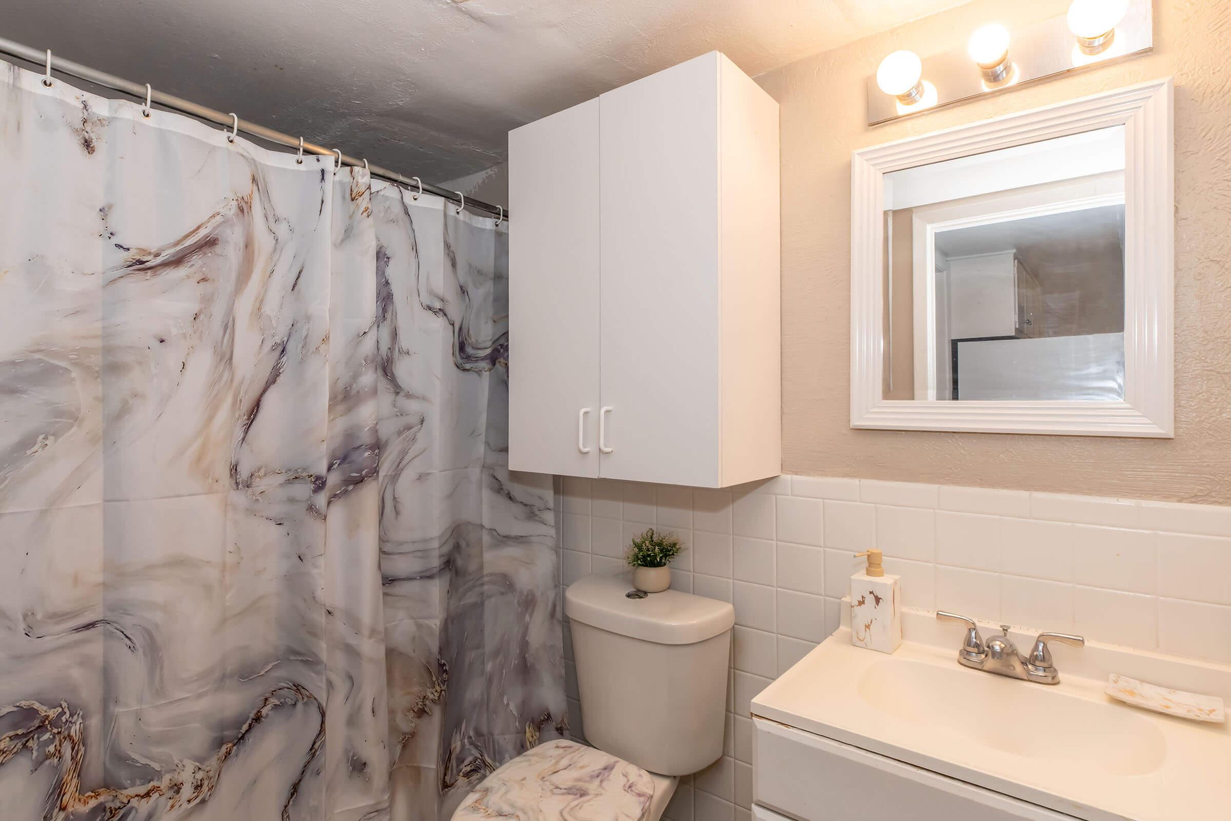 A modern bathroom featuring a marble-patterned shower curtain, a white vanity with a sink, and a compact toilet. There is a small cabinet above the toilet and a mirror with a white frame. Soft lighting is provided by three bulbs above the mirror, and a decorative plant adds a touch of greenery.