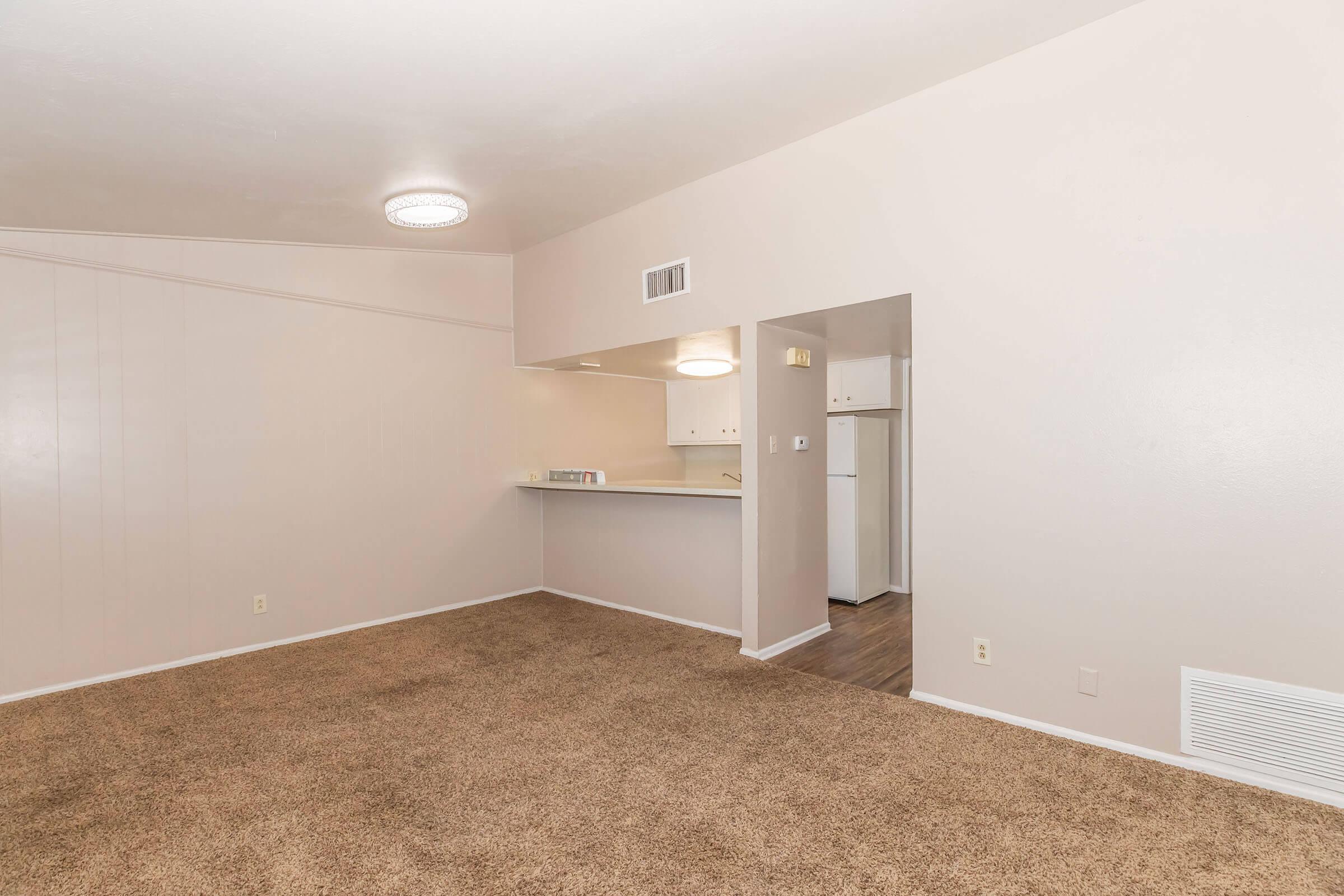 Spacious living area with carpeted floor, neutral wall colors, and ceiling lighting. A doorway leads to a small kitchen area featuring a bar counter and white appliances, including a refrigerator. Enhanced natural light and an open layout create a welcoming environment.