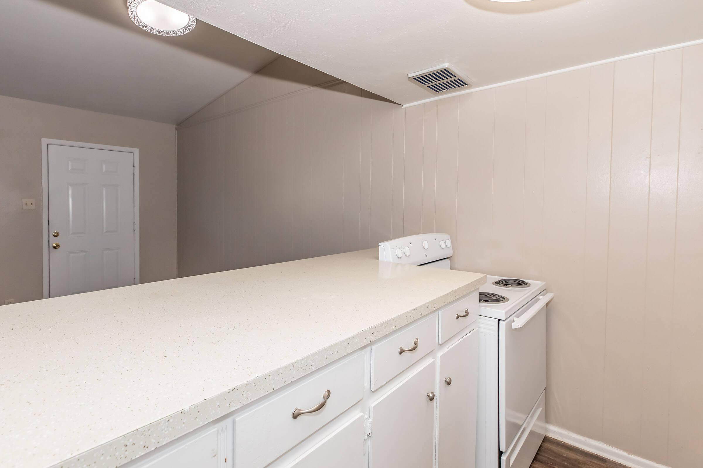 A clean, modern kitchen with a white countertop and an electric stovetop. The kitchen features white cabinets and a white stove, with a door visible in the background. Soft lighting illuminates the space, creating a bright and inviting atmosphere.