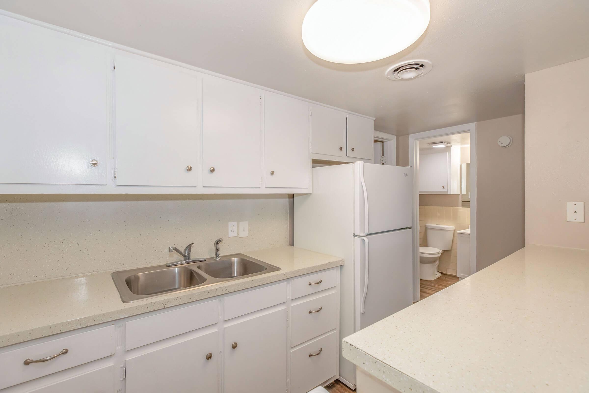 A clean, modern kitchen featuring white cabinetry, a double sink, and a refrigerator. The countertop is light-colored, and the space has neutral walls. In the background, a doorway leads to a bathroom. The area is well-lit with a ceiling light fixture.