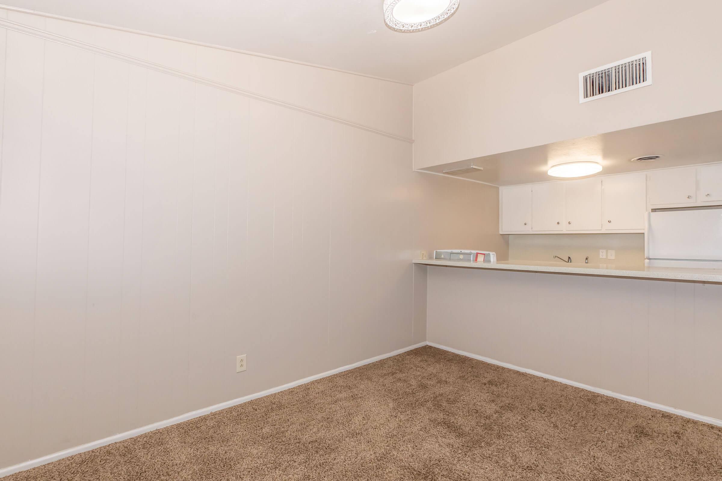 A spacious room with beige walls and carpet, featuring a small kitchen area with light-colored cabinets and a countertop. An overhead light fixture is visible, and there is an empty wall space suitable for furniture or decor. The kitchen is partially visible in the background, showing a clean and organized space.