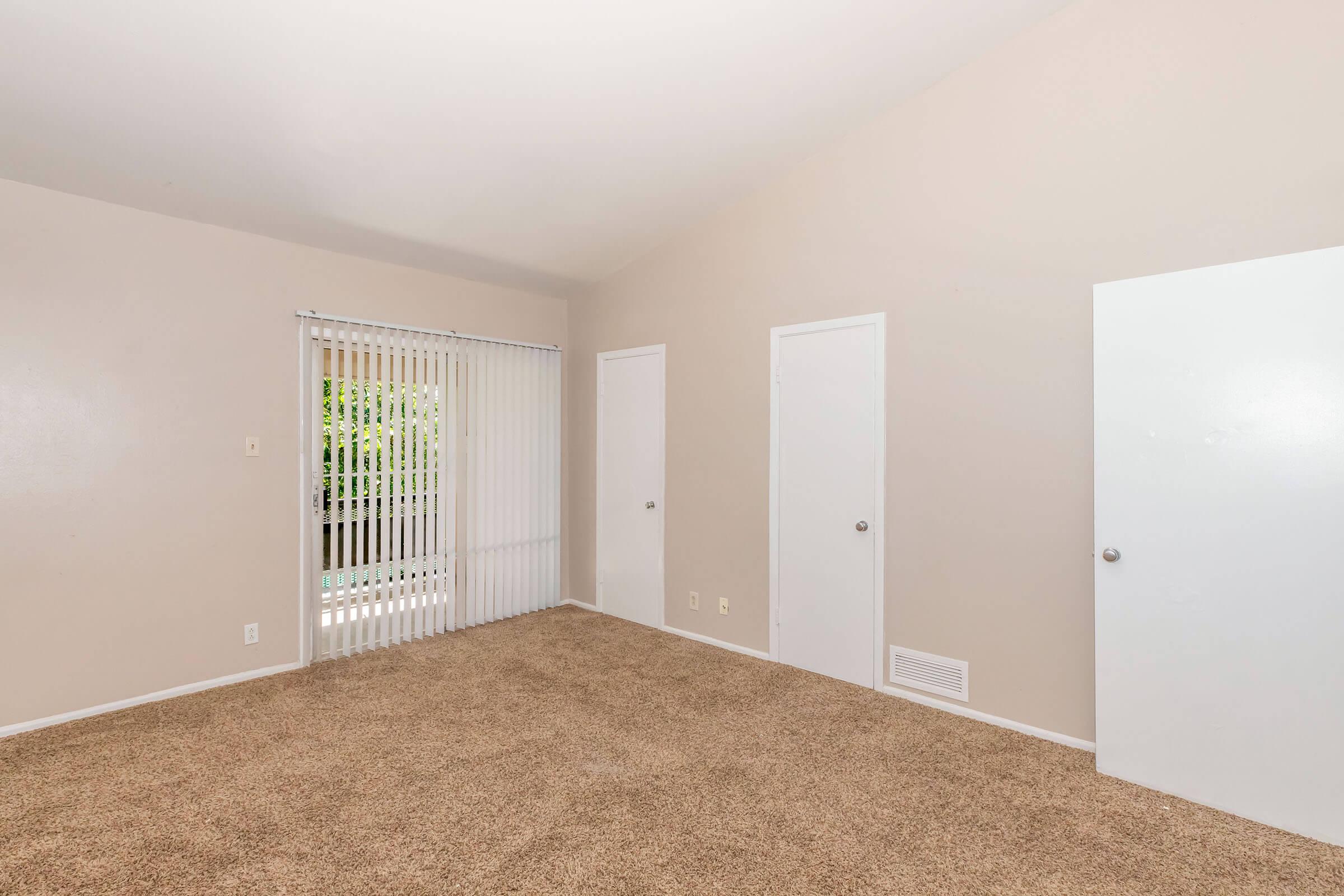 A spacious, empty room with light beige walls and plush carpet. There are two white doors on one wall, a window covered with vertical blinds letting in natural light, and a vent on the floor. The ceiling has a gentle slope, providing an open and airy feel.