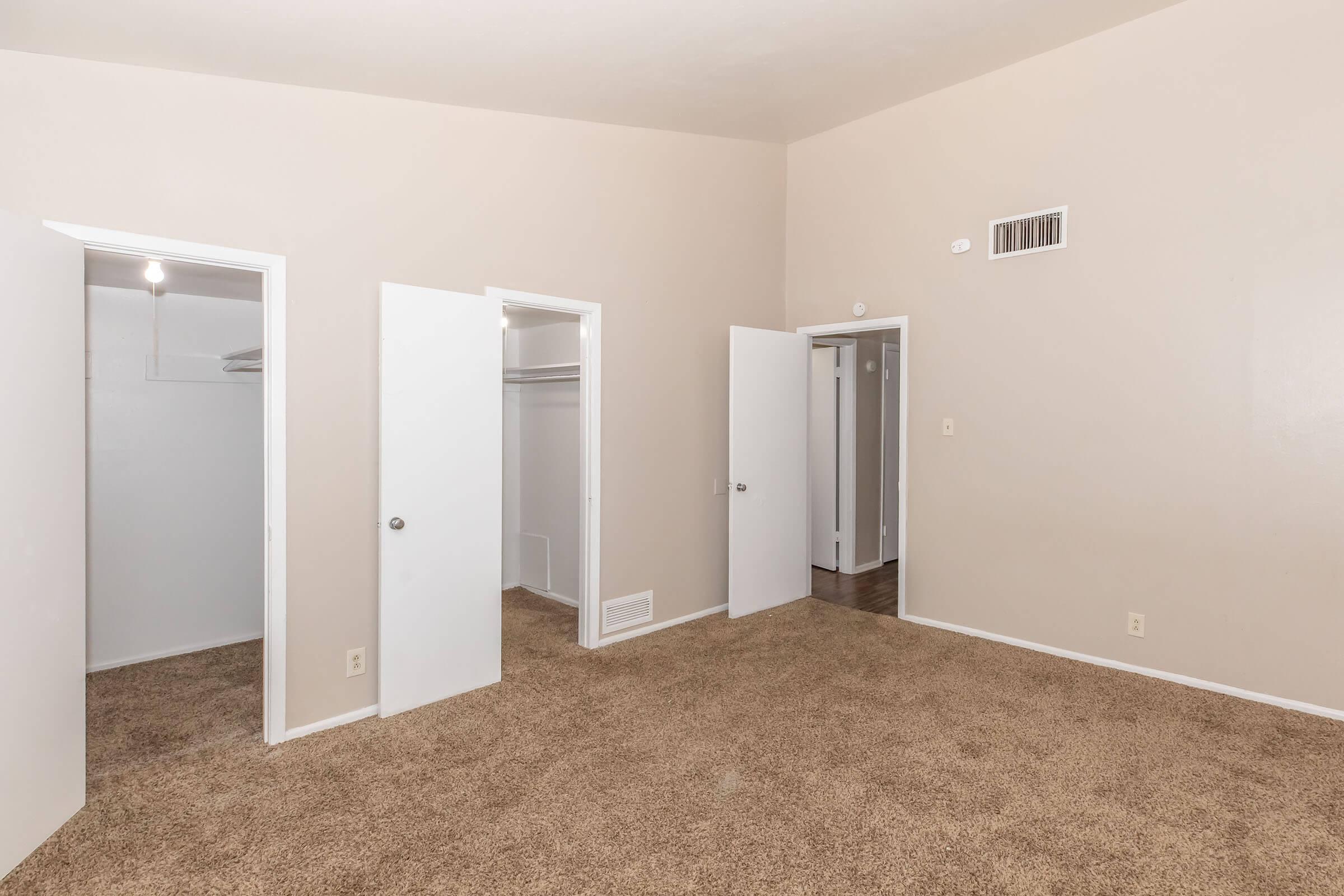 Interior of a room featuring beige walls and carpet. Three white doors are visible: one leading to a closet, another to an unknown space, and a third door. The room has a high ceiling and a small vent on the wall, with natural light illuminating the space.