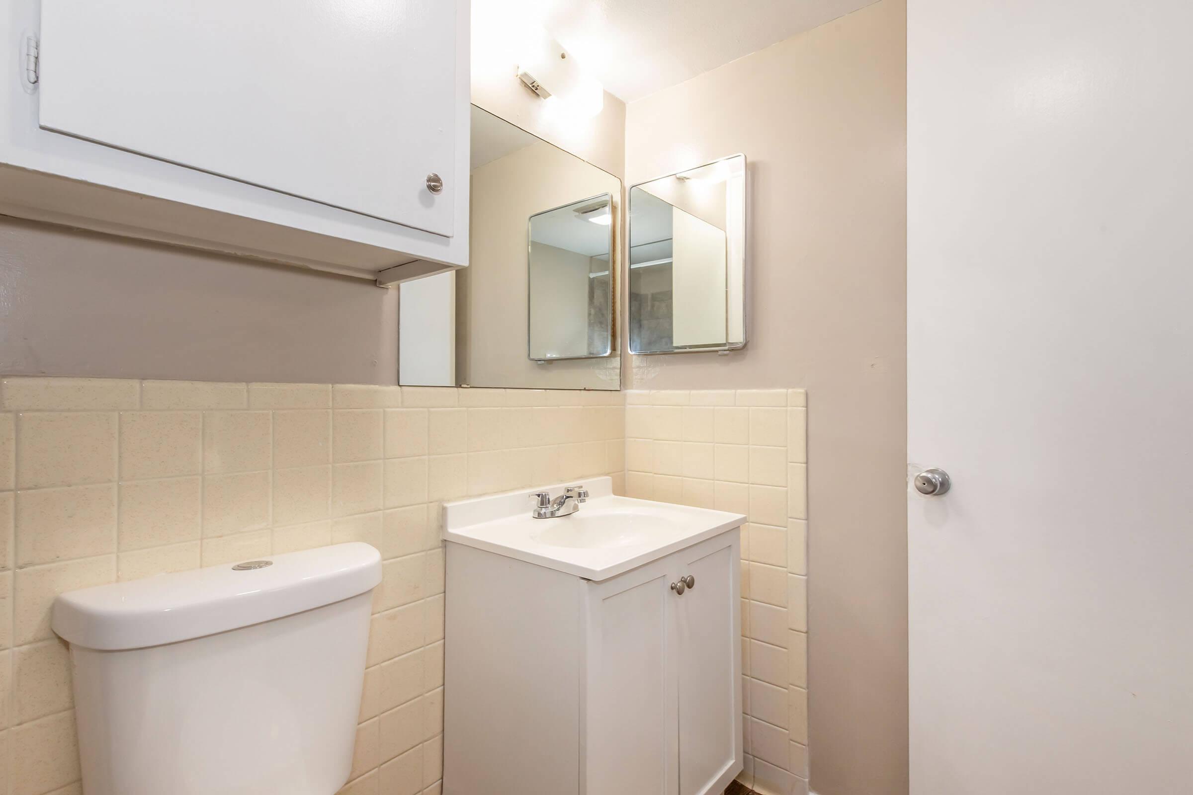 A small bathroom featuring a white toilet, a white sink with a cabinet beneath, and two mirrors above the sink. The walls are painted light beige, and the tiled backsplash is a light yellow. A door on the right leads to another space. The overall decor is simple and functional.