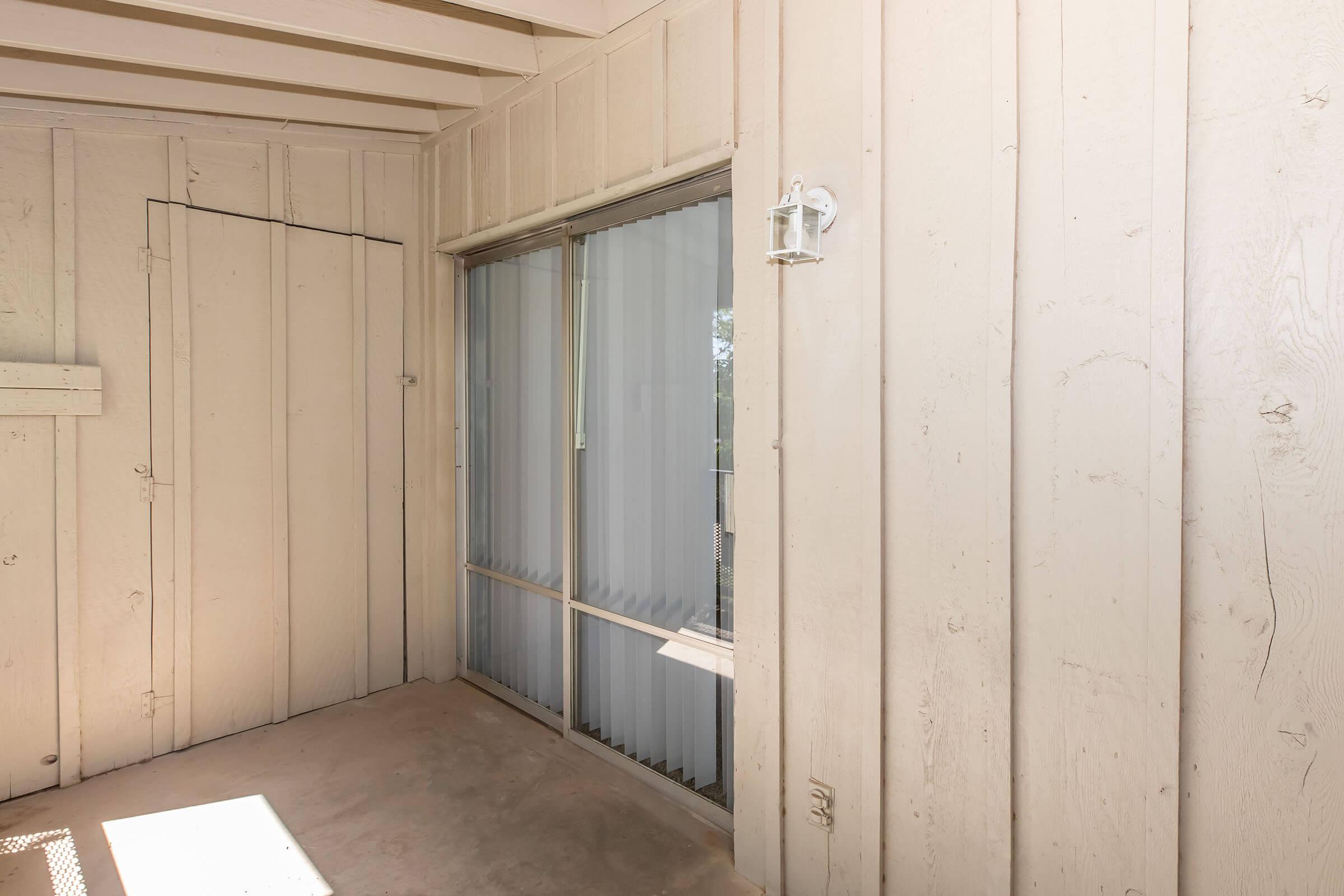 An empty, partially enclosed porch with beige wooden walls and a large window featuring vertical blinds. The space has a concrete floor and a door beside the window, creating a simple, minimalistic atmosphere with natural light coming in.