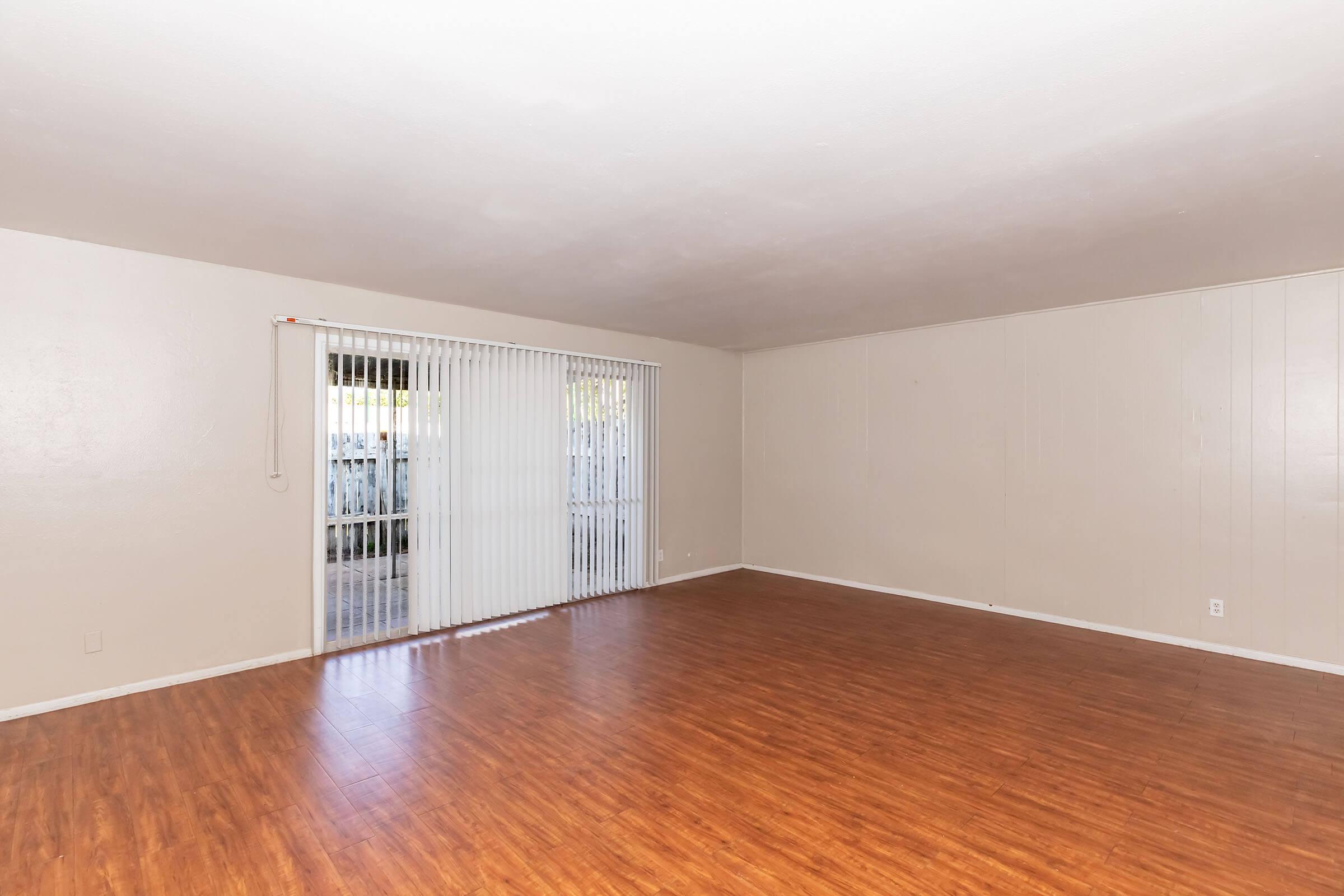 Empty room with light-colored walls and a wooden floor. A large sliding glass door with vertical blinds allows natural light to enter from outside. The space is uncluttered, creating a bright and open atmosphere.