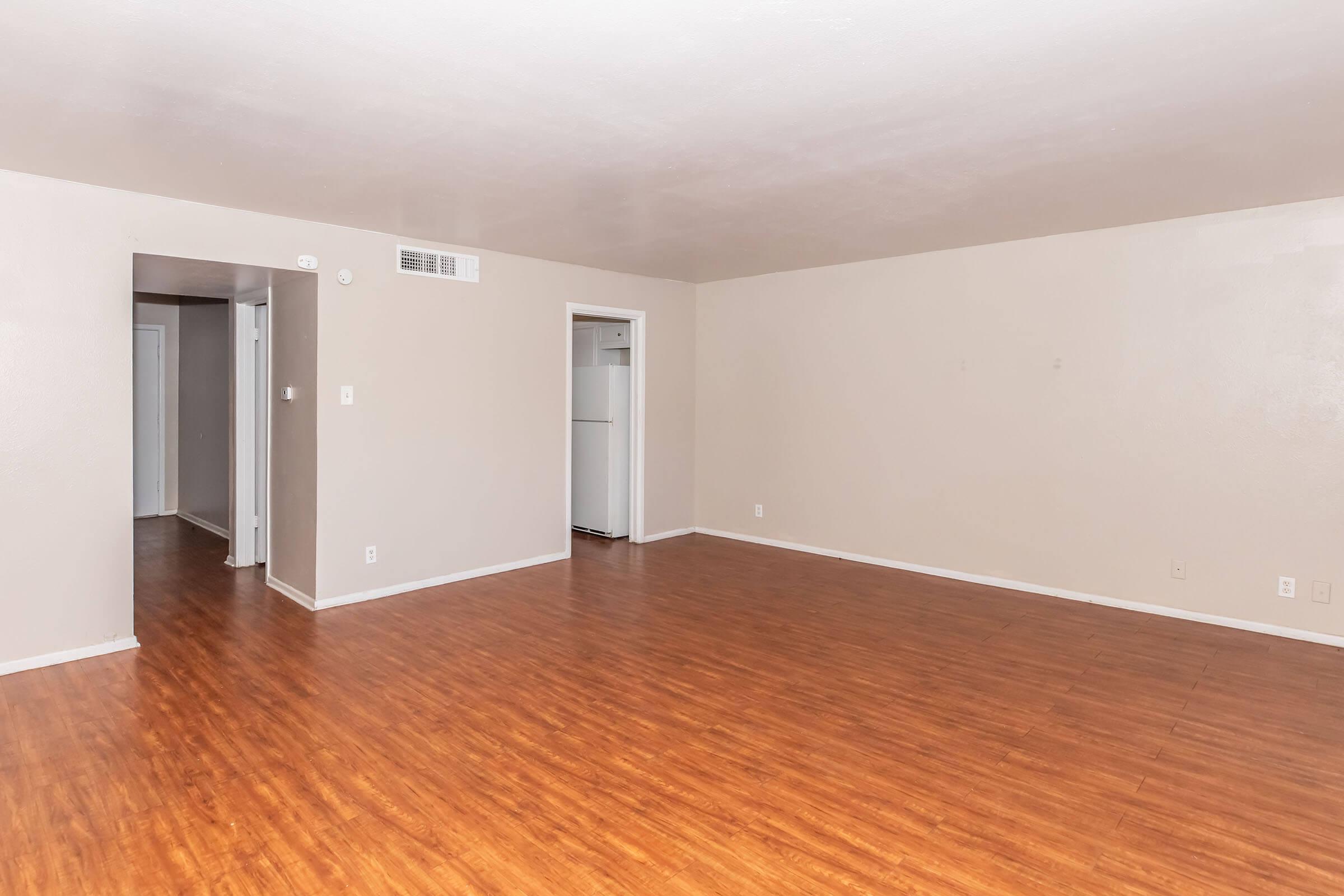 A spacious, empty living room with light beige walls and a medium brown laminate floor. There are two doorways leading to other areas, and a refrigerator is visible through one doorway. The room is well-lit and uncluttered, creating a simple and airy atmosphere.