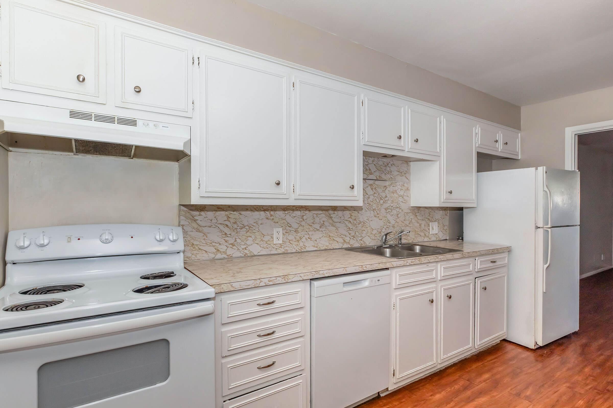 A modern kitchen featuring white cabinets, a marble backsplash, a white stove and oven, and a white refrigerator. The countertops are light-colored, and the flooring is wood. The kitchen has an open layout with a doorway leading to another room visible in the background.