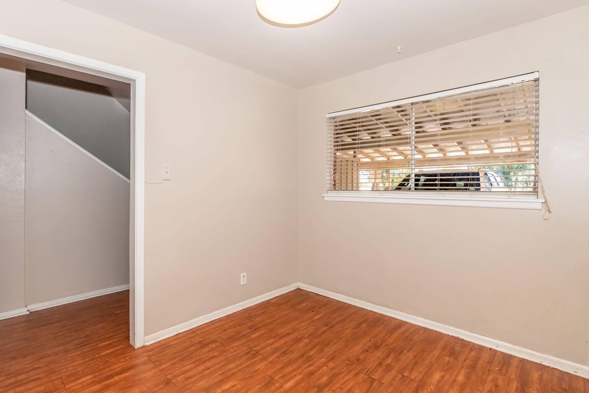 A small, empty room with light-colored walls and a wooden floor. There is a window with blinds letting in natural light, and a staircase is visible in an adjacent hallway. The room appears clean and unfurnished, suitable for various purposes.