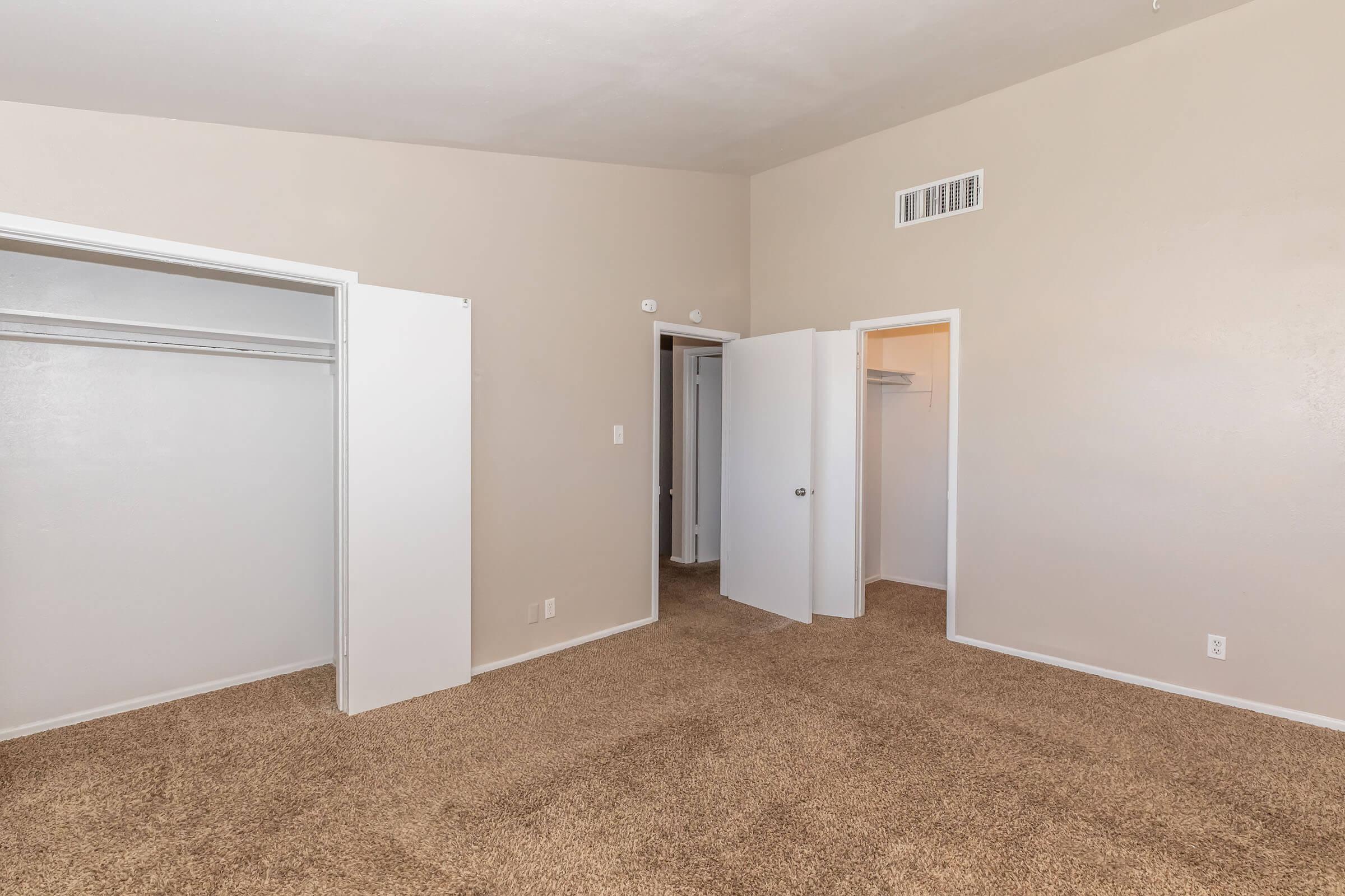 A spacious, empty room with light beige walls and a carpeted floor. There are two open closet doors on the left, and a doorway leading to another room is visible in the background. The overall lighting is bright, creating an airy atmosphere.