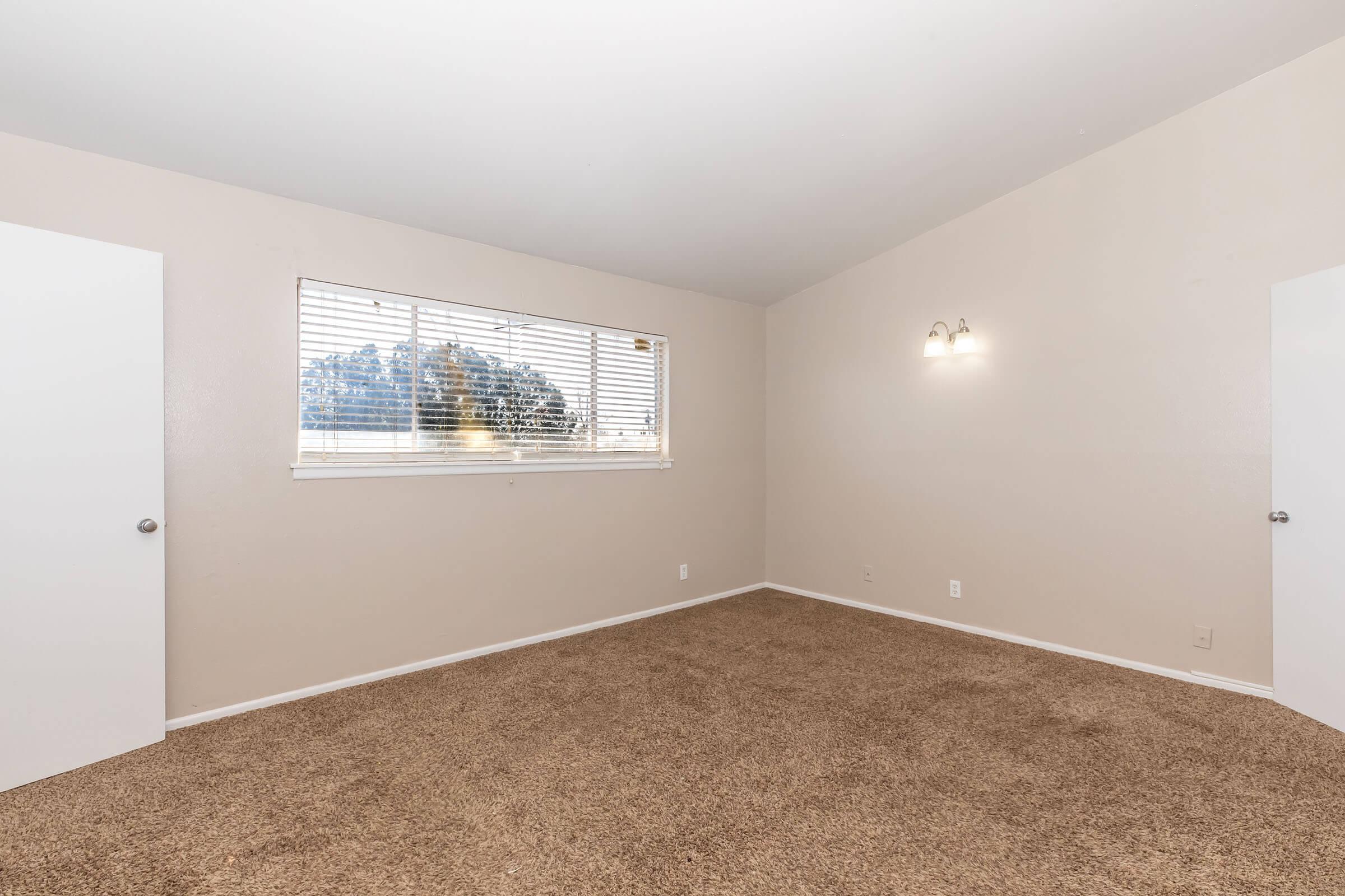 A spacious, empty room featuring light beige walls and a carpeted floor. There are two white doors on either side and a window with blinds, allowing natural light to enter. Wall sconces provide additional lighting, creating a bright and inviting atmosphere. Ideal for various uses such as a bedroom or office space.
