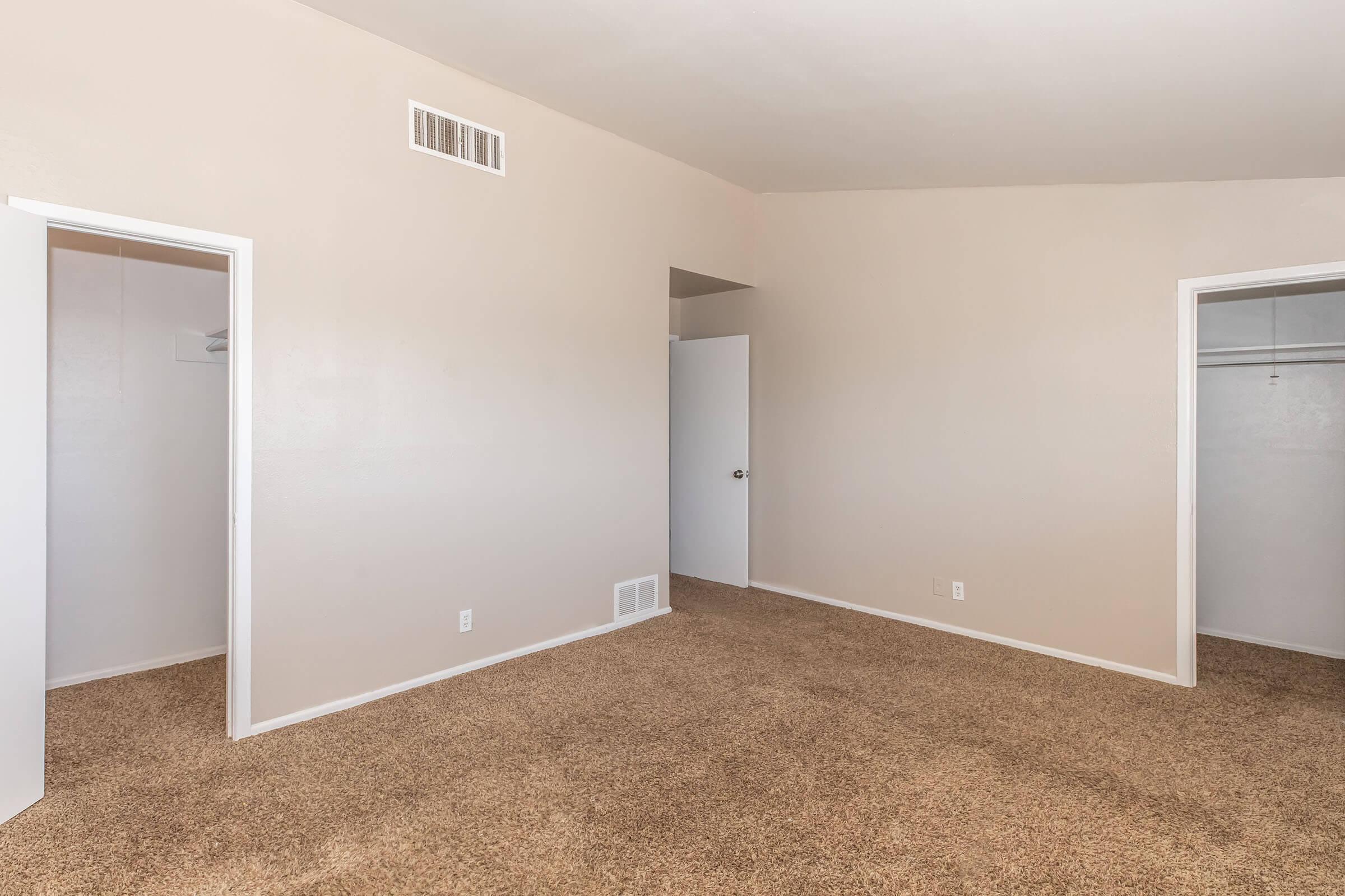 A spacious, empty room with beige walls and light brown carpet. There are two doorways on the left leading to closets, and a third door on the right. The room is well-lit and features a simple and clean design, suitable for various purposes.
