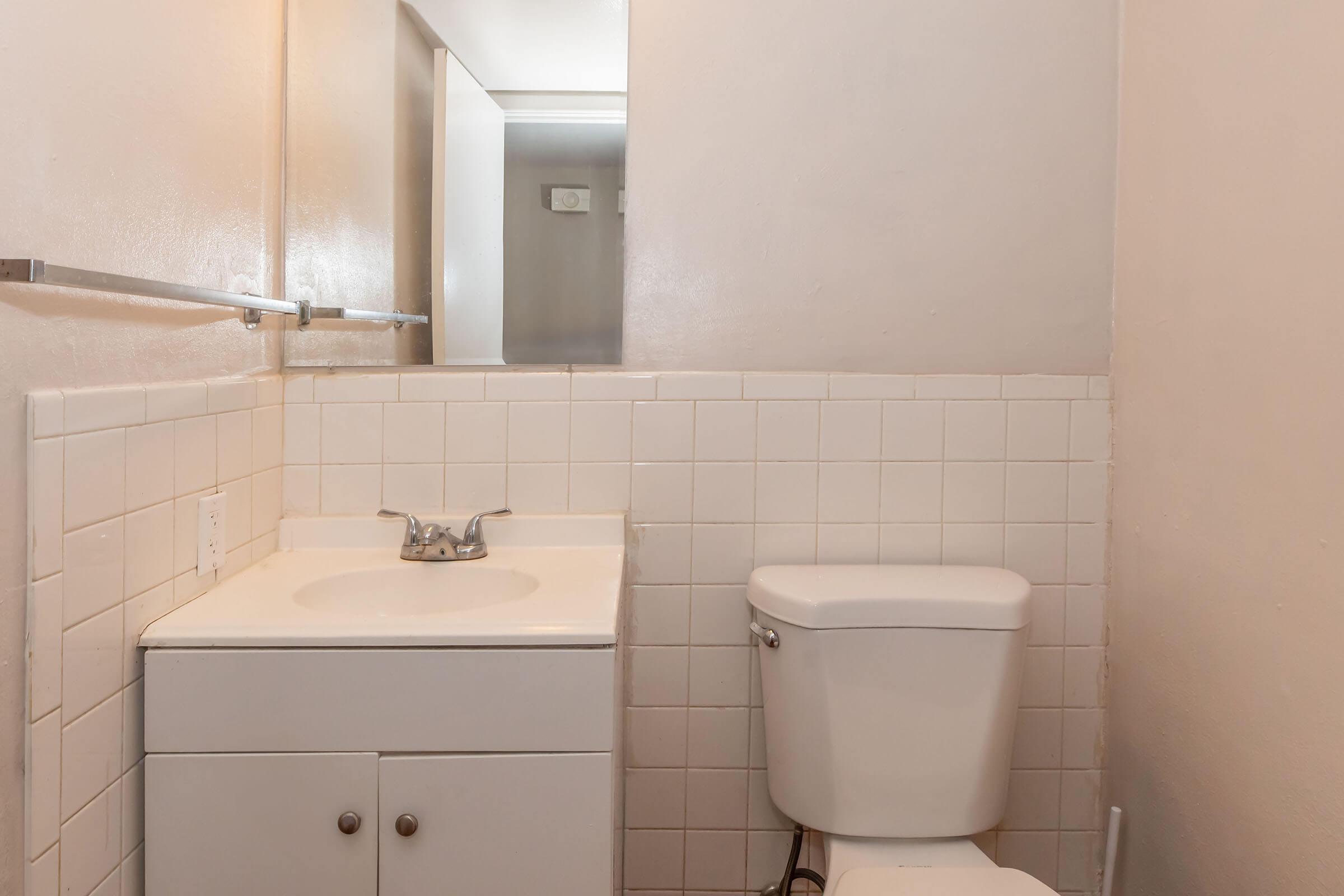 A small bathroom featuring a white sink with a single faucet and a white toilet. The walls are painted a light color, with tiled portions on the lower half. A mirror is visible in the background, reflecting light into the space.