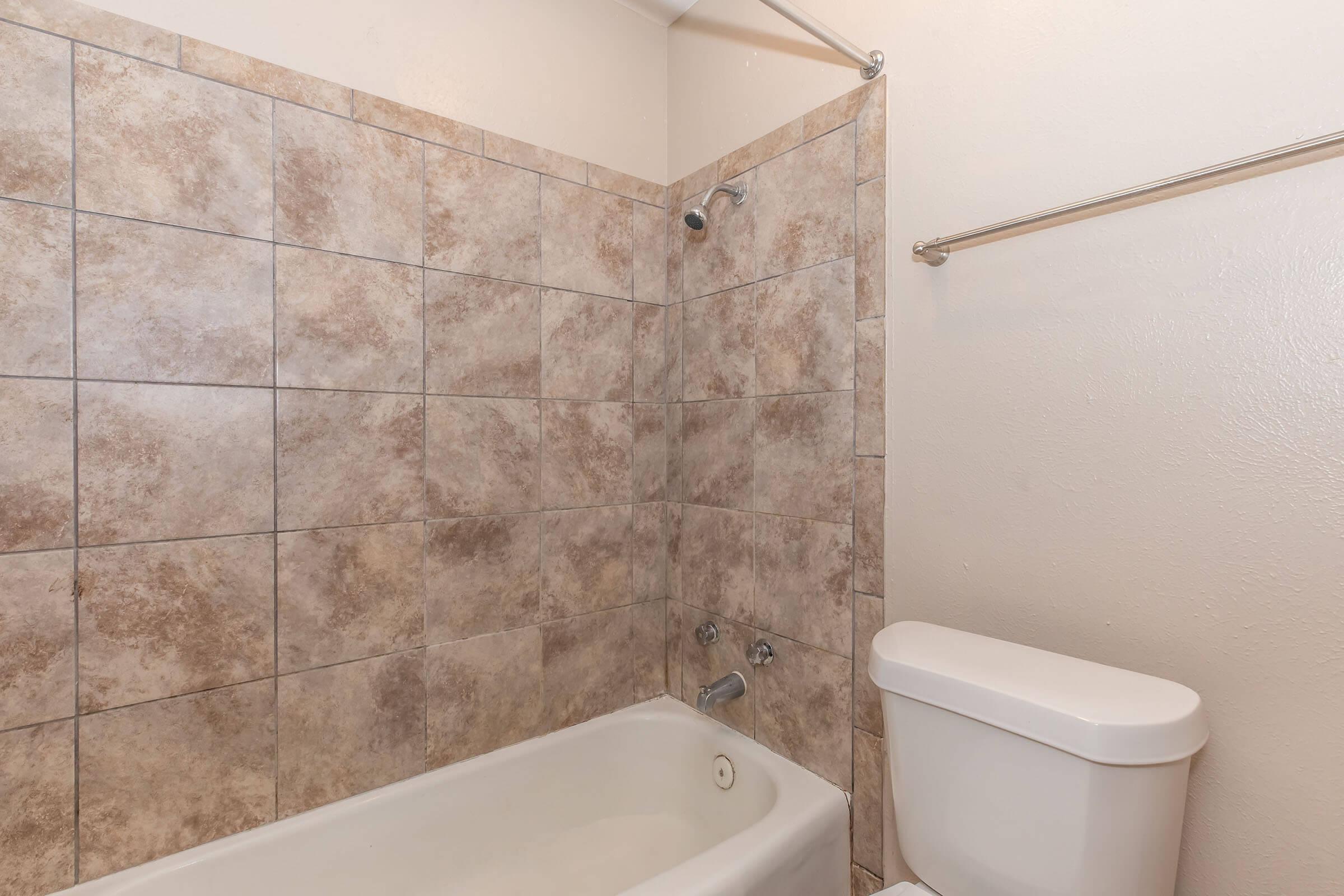 Bathroom featuring a tiled wall, with a bathtub on the left side and a toilet on the right. The tiles have a neutral, earthy tone, and a showerhead is mounted above the tub. There is also a towel bar installed on the wall. The overall space appears clean and well-maintained.