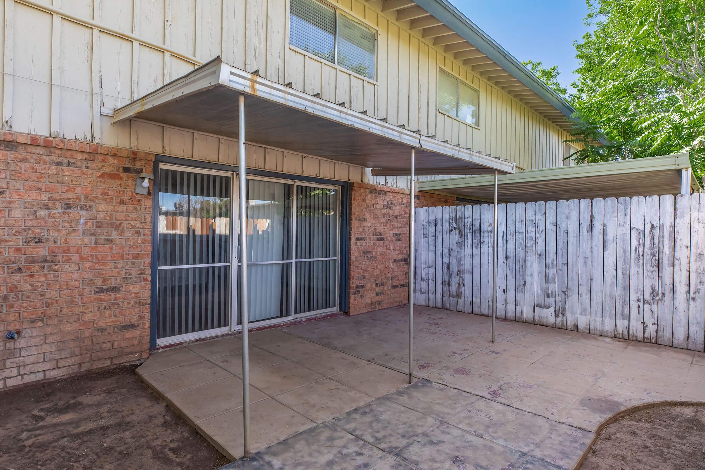 A spacious outdoor patio area with a concrete floor, covered by a metal awning. The background features a brick wall of a house, sliding glass doors, and a white wooden fence. Lush greenery can be seen in the distance, creating a serene outdoor setting.