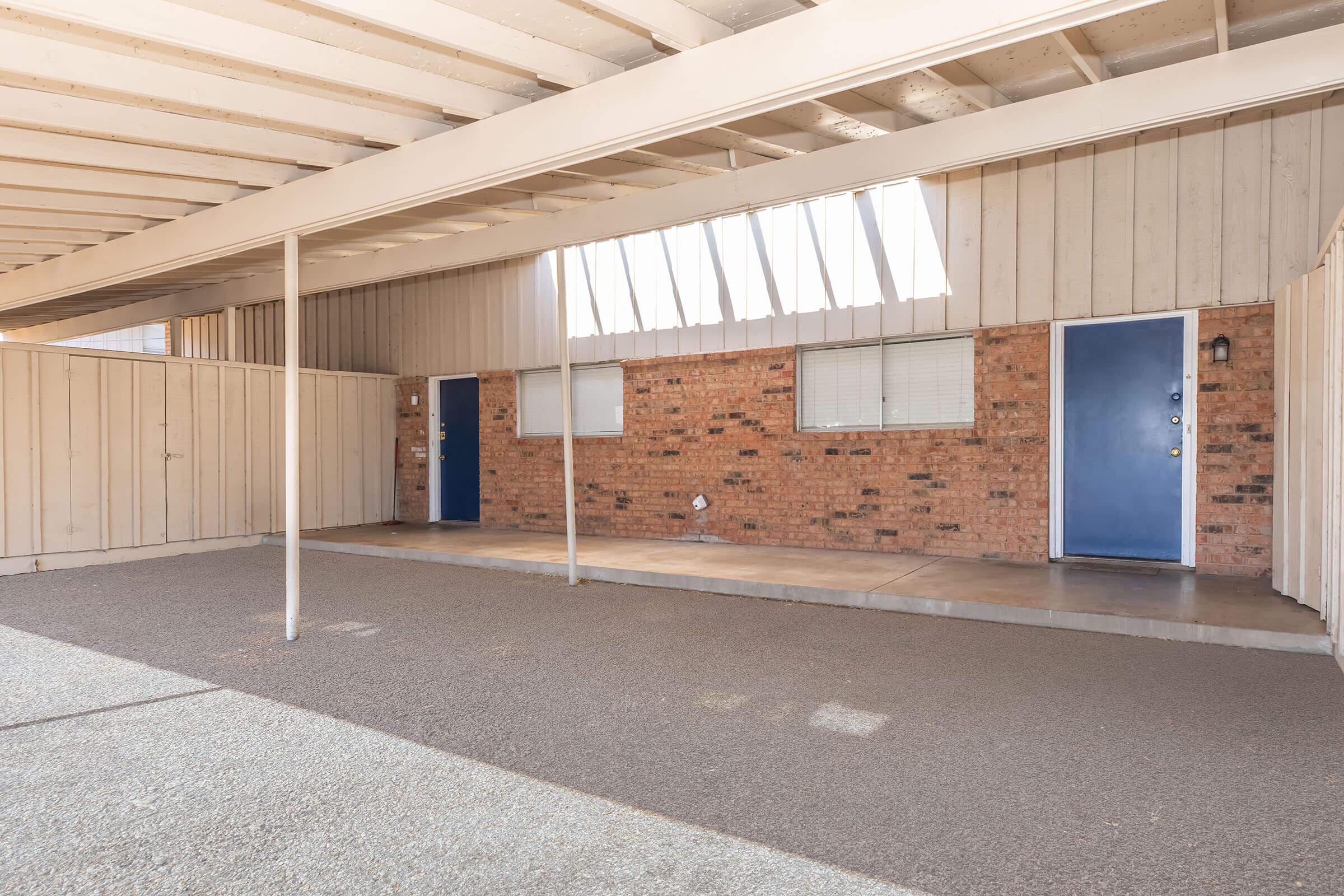 Two blue entrance doors situated in a brick wall, framed by a spacious covered area with a concrete floor. The roof has exposed beams, and there are windows above the doors allowing natural light. The surrounding walls are textured and painted in a neutral color, creating a welcoming exterior atmosphere.