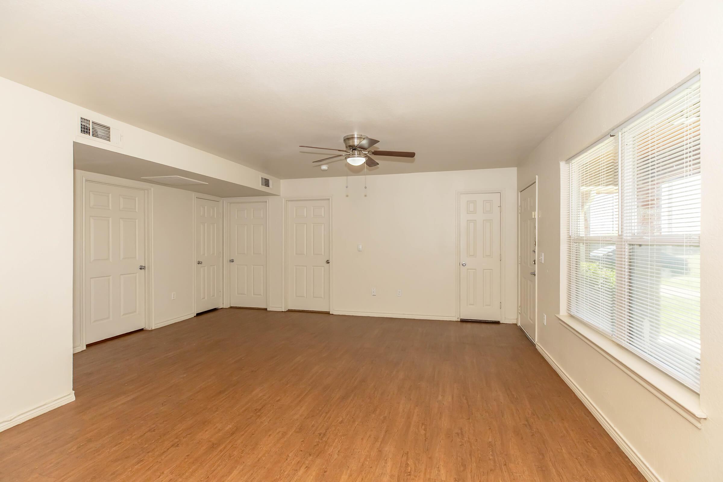 Spacious living area with light-colored walls, wooden flooring, and a ceiling fan. There are three closed doors along one wall, and a large window with blinds allowing natural light to enter the room. The overall atmosphere is bright and inviting, perfect for furniture arrangement and decoration.
