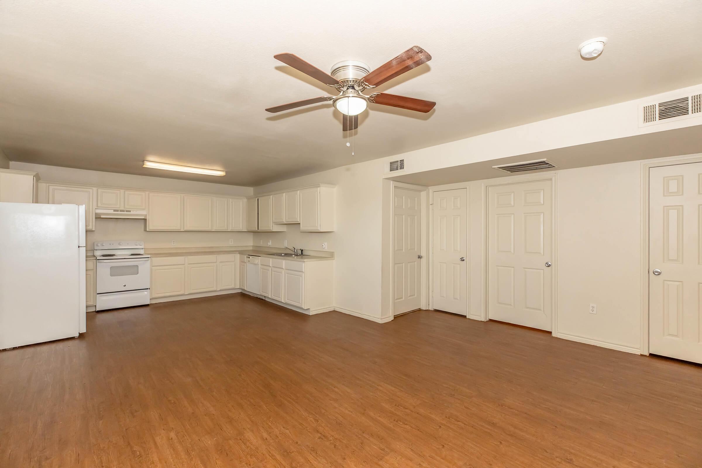 Spacious, well-lit apartment interior featuring a living area with wooden flooring, ceiling fan, and a kitchen area. The kitchen includes white cabinets, an oven, and a refrigerator. Multiple doors lead to storage or closet spaces, creating a functional and open layout.