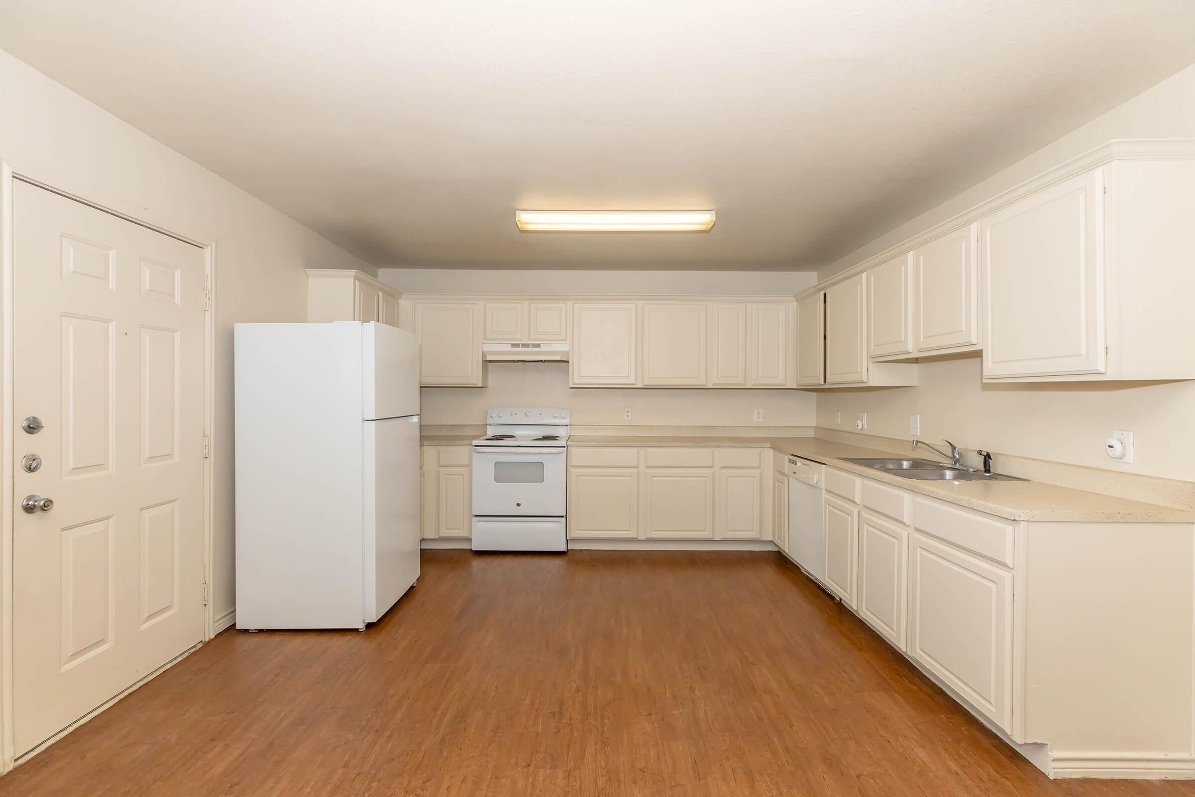 Spacious kitchen featuring white cabinetry, a white refrigerator, a stove, and ample countertop space. The room has a light wood floor and large windows, providing natural light. The layout includes a door leading outside, contributing to an open and inviting atmosphere.