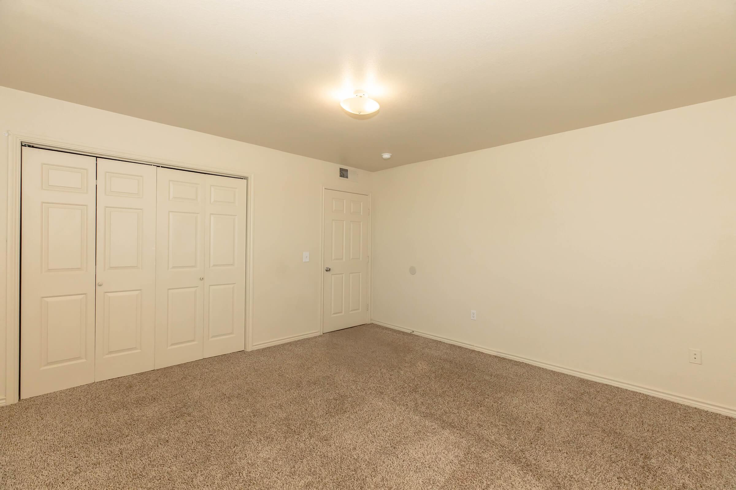 A spacious, empty room with light beige walls and soft carpet flooring. There are two sets of double doors, likely closets, on one side, and a single door on the opposite side. A ceiling light provides illumination, highlighting the room's neutrality and readiness for personalization.
