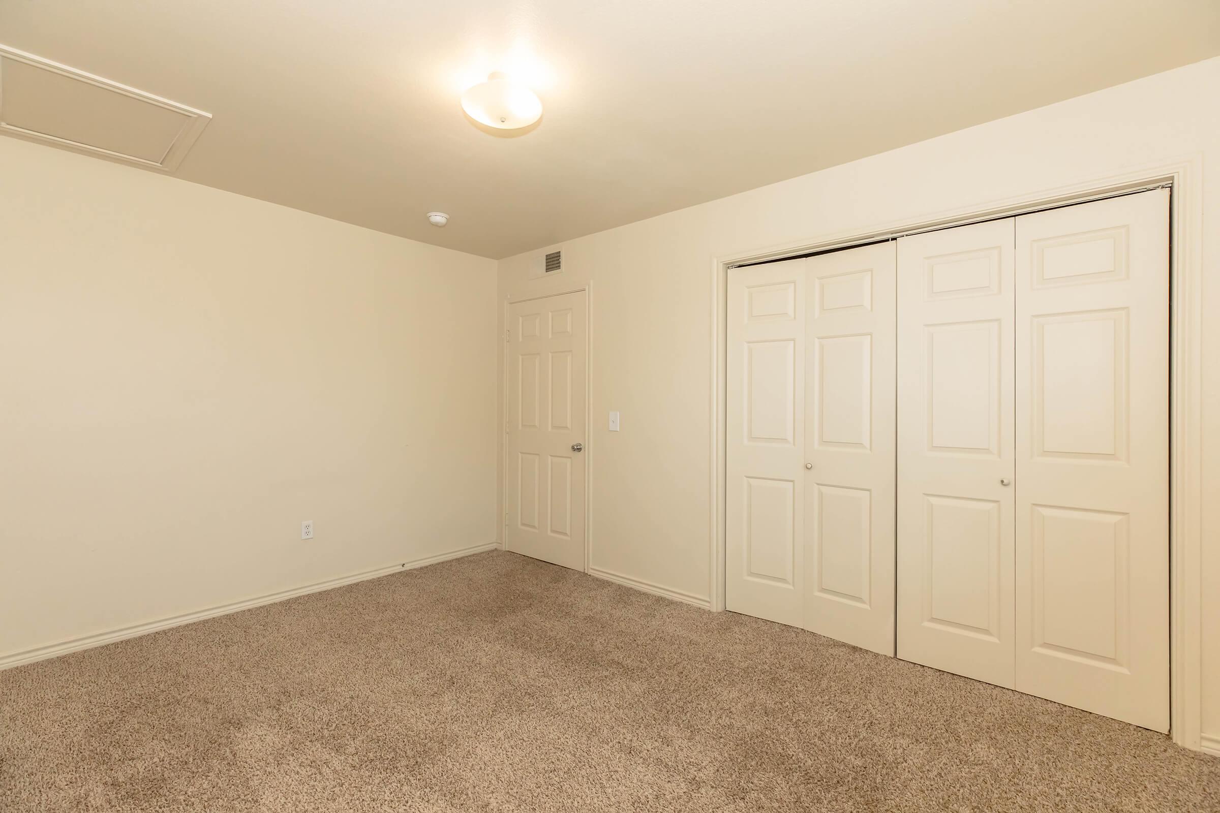 A clean, empty room with beige walls and carpeted flooring. There is a ceiling light fixture, and two sets of double doors leading to closets are visible on one wall. The room appears spacious and well-lit, suitable for various purposes.