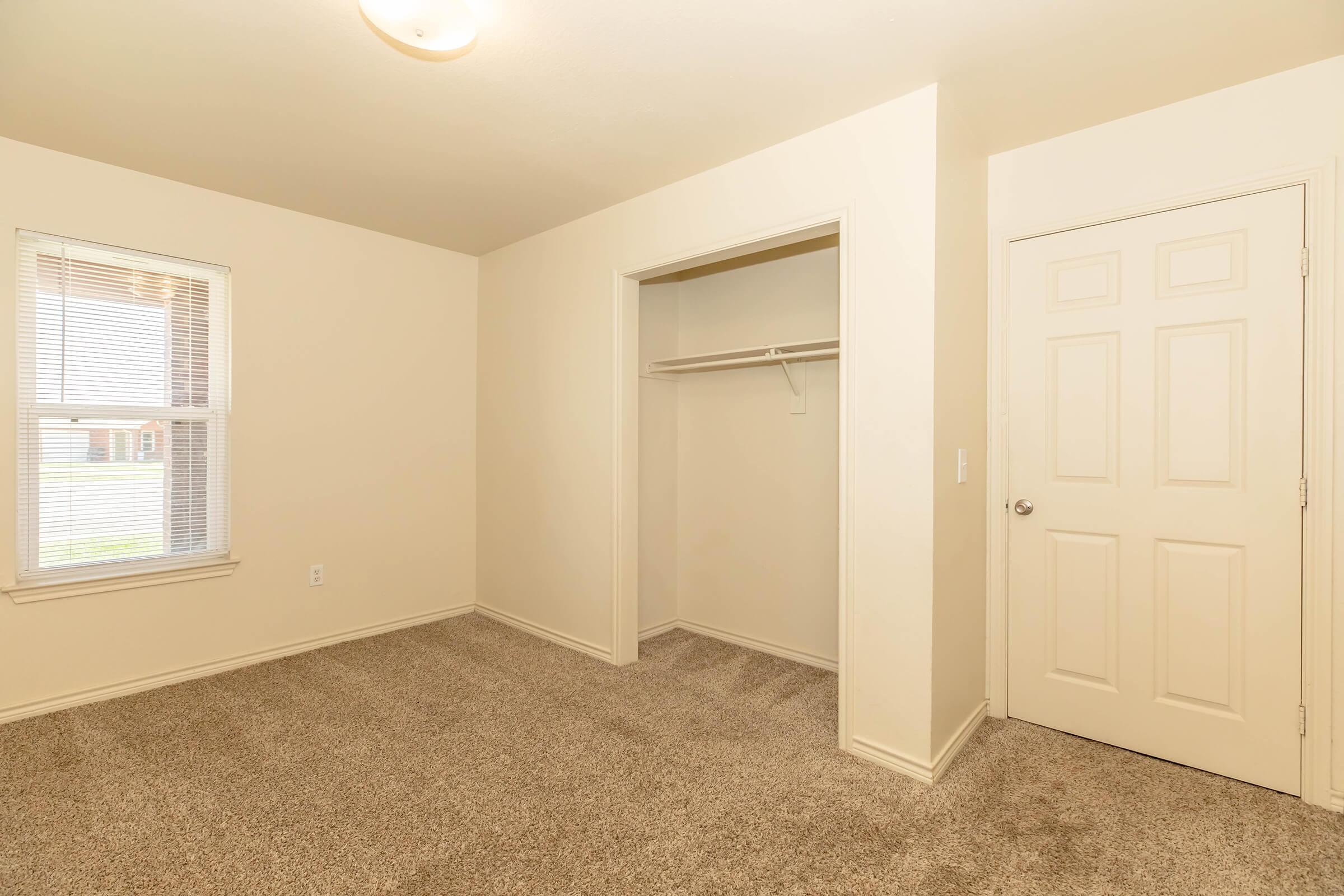 A view of an empty bedroom with light beige walls and carpet, featuring a window with blinds, an open closet with a hanging rod, and a door leading to another room. The space is well-lit and has a simple, clean design, creating a cozy and inviting atmosphere.