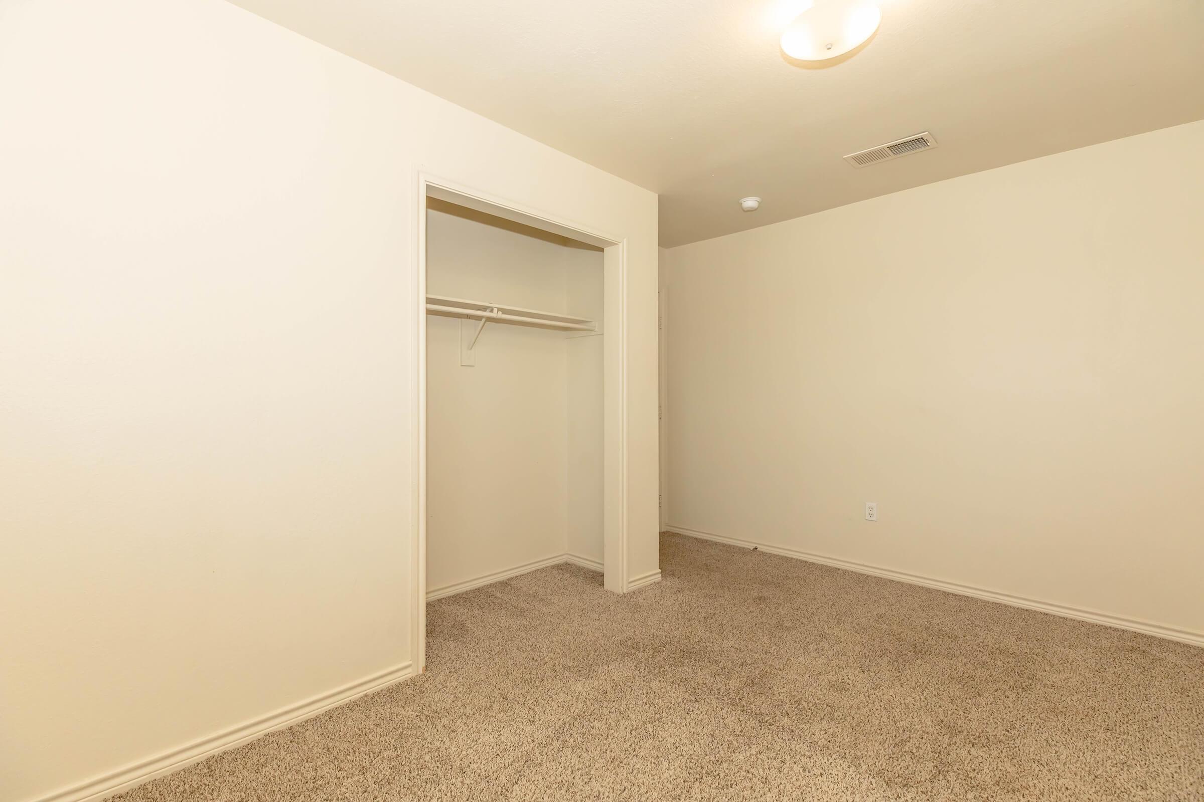 Empty room with beige walls and carpeted floor. There is a small closet on one side with a shelf and hanging rod. A ceiling light fixture provides illumination. The room is unoccupied, creating an open and spacious atmosphere.