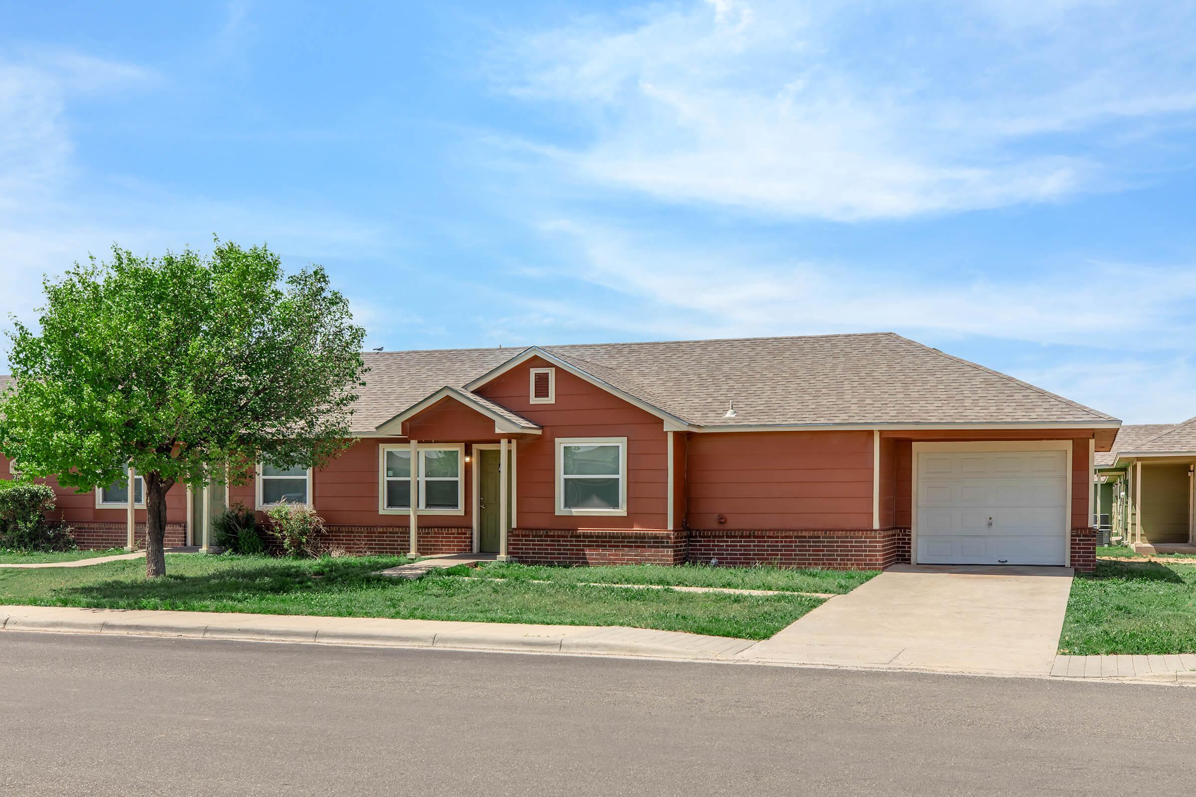 A single-story house with a reddish-brown exterior, featuring a front porch and a garage. The lawn is well-maintained, with a tree and shrubs in the front yard. The sky is clear with a few clouds, and the street is paved.