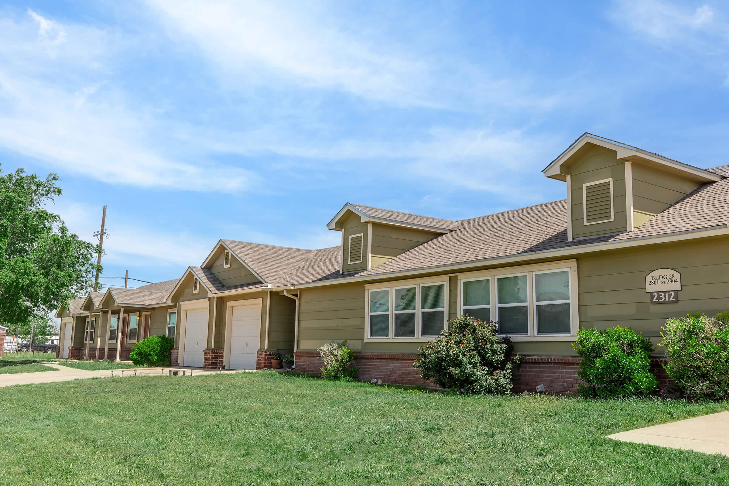 A row of single-story houses with light brown siding and gabled roofs, situated on a green lawn. The sky is blue with scattered clouds. A driveway leads to one of the houses, and small shrubs are planted near the foundation. The scene conveys a calm and inviting residential neighborhood atmosphere.