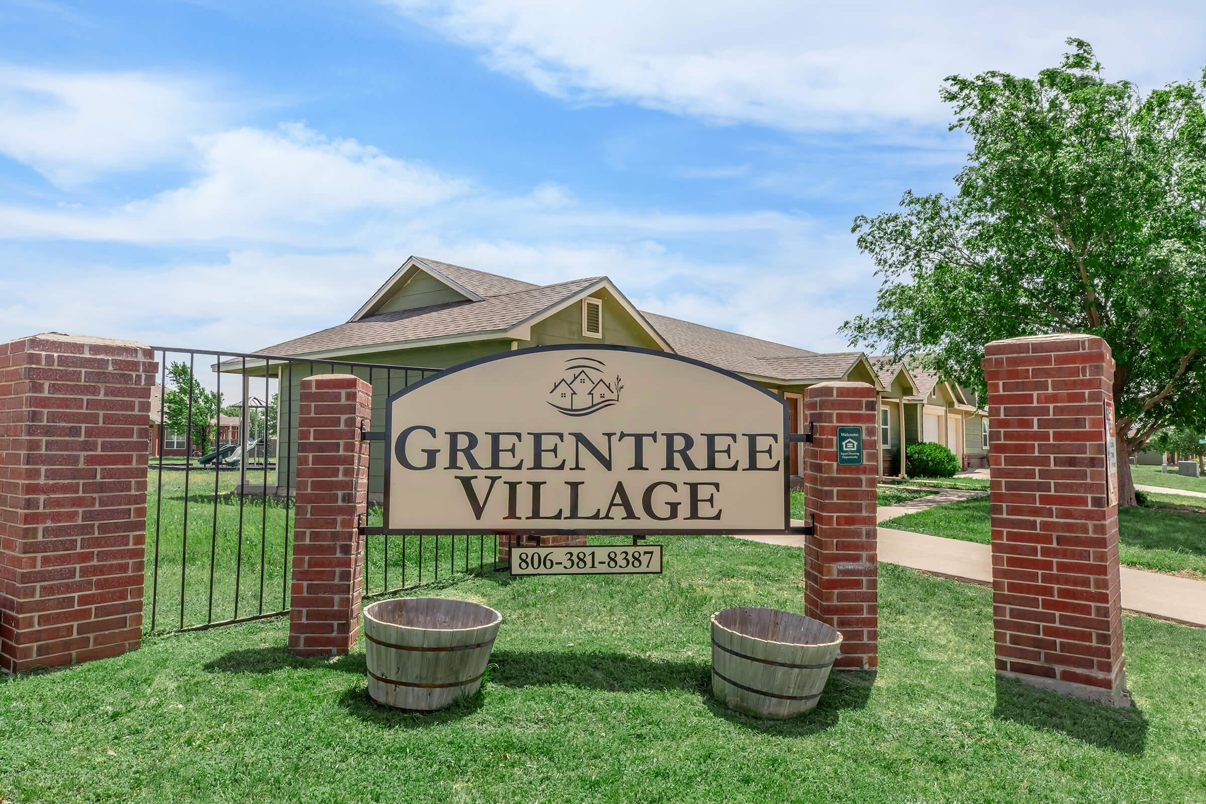 Sign for Greentree Village displayed on brick columns, surrounded by green grass and trees, with a clear blue sky in the background. The sign includes a phone number, suggesting it may be an apartment or community complex.