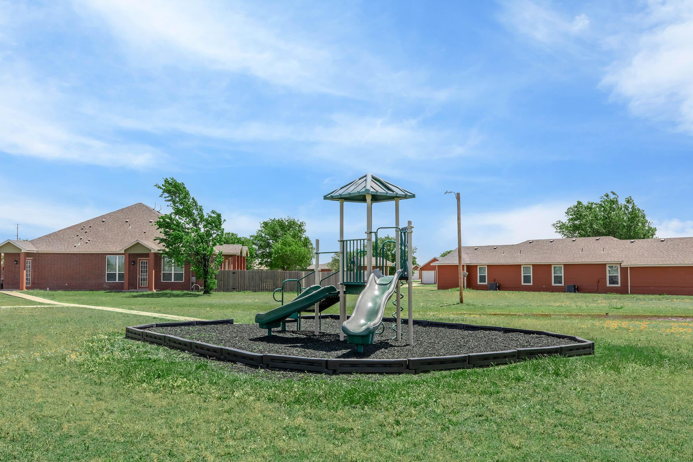 A playground featuring two slides, one green and one silver, situated on a grassy area surrounded by a black rubber border. In the background, there are two residential buildings with sandy-colored exteriors under a bright blue sky with scattered clouds.