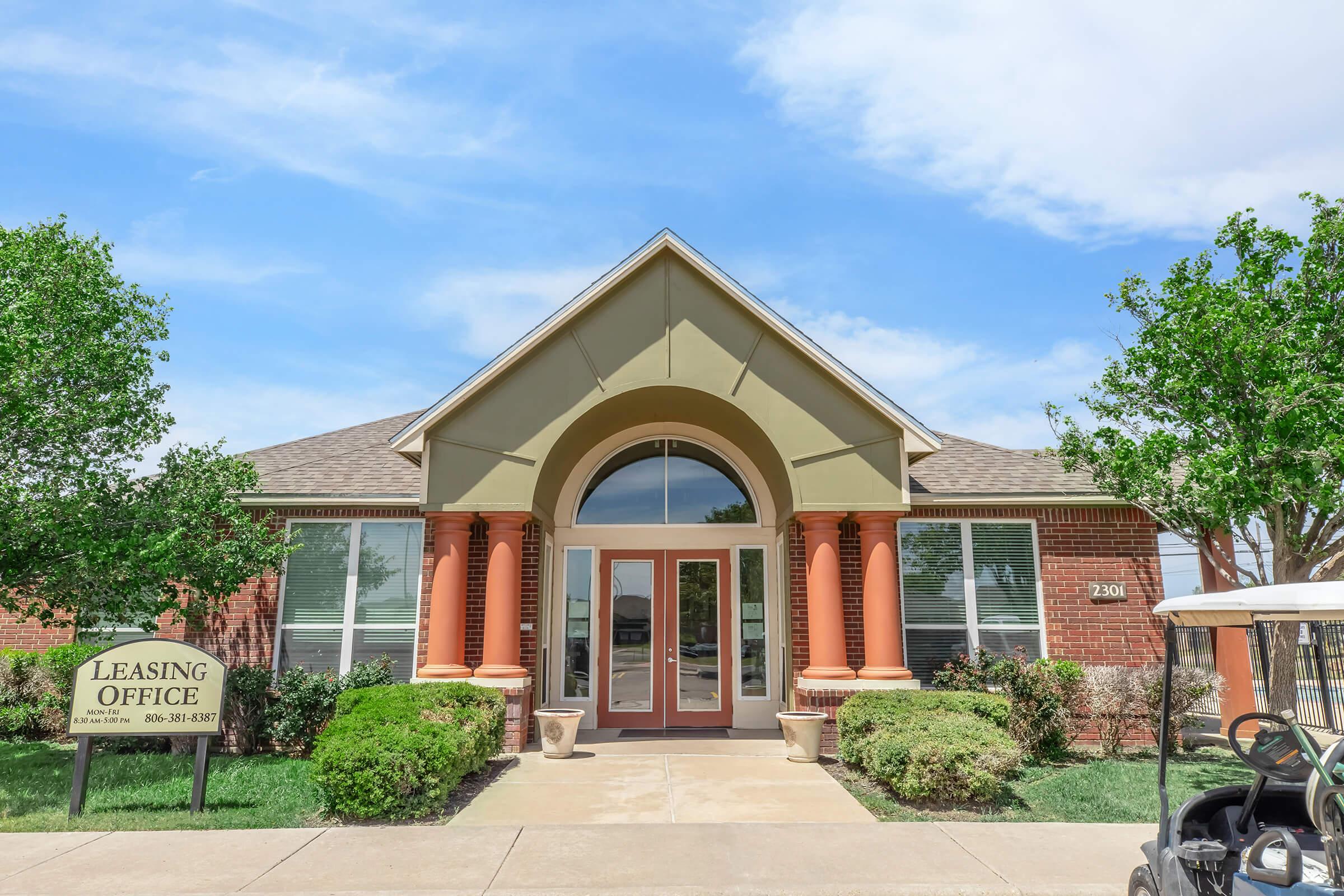 A modern leasing office building with a peaked roof, featuring large front doors and decorative pillars. The exterior has a mix of brick and green accents, surrounded by small bushes and trees. A sign in front indicates it's the leasing office. The sky is clear with a few clouds.