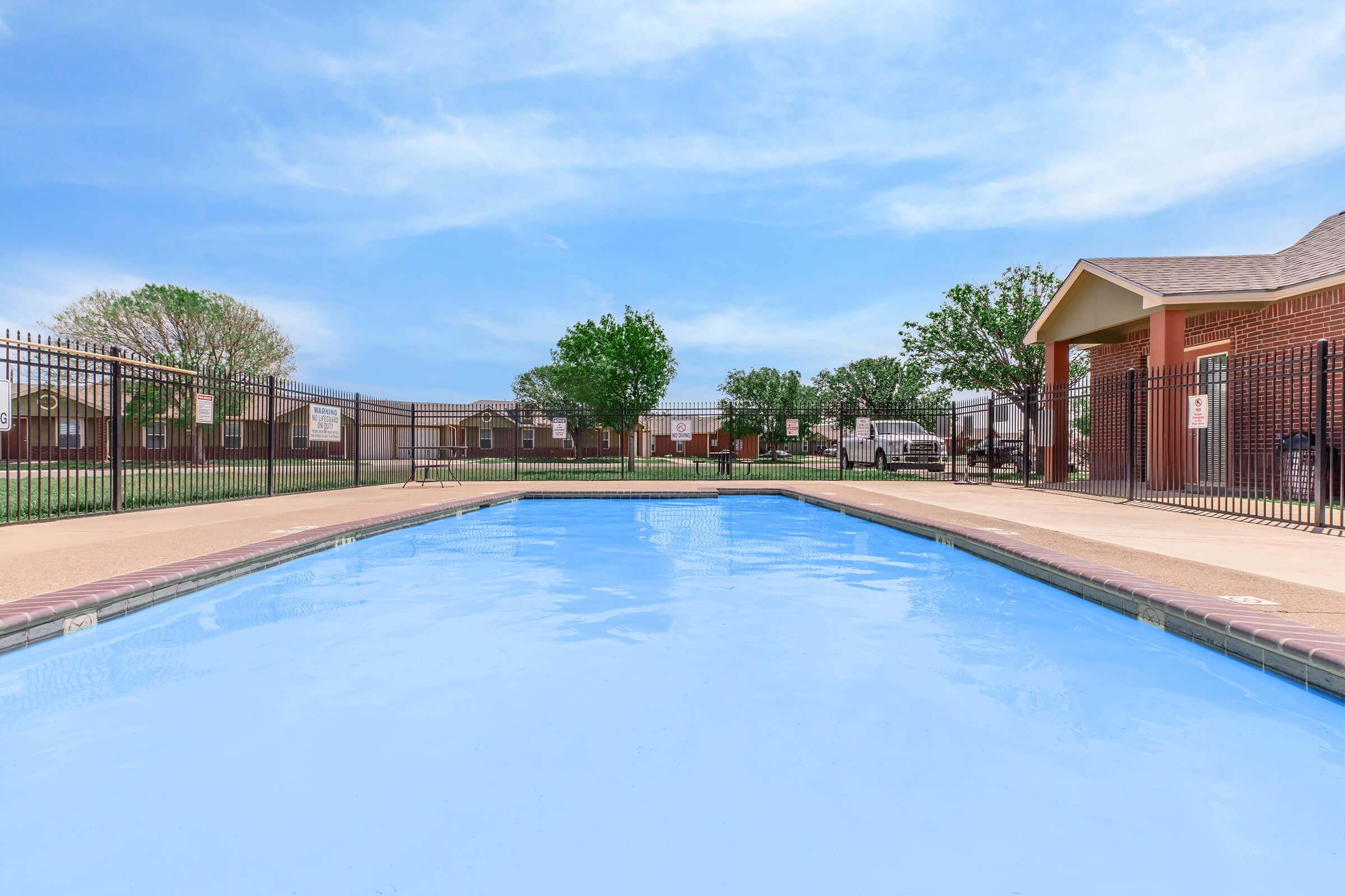 A clear blue swimming pool with a smooth surface, surrounded by a well-maintained patio and a fence. Lush green trees and grass are visible in the background, along with a few buildings. The sky is bright blue with some clouds, creating a peaceful outdoor atmosphere.