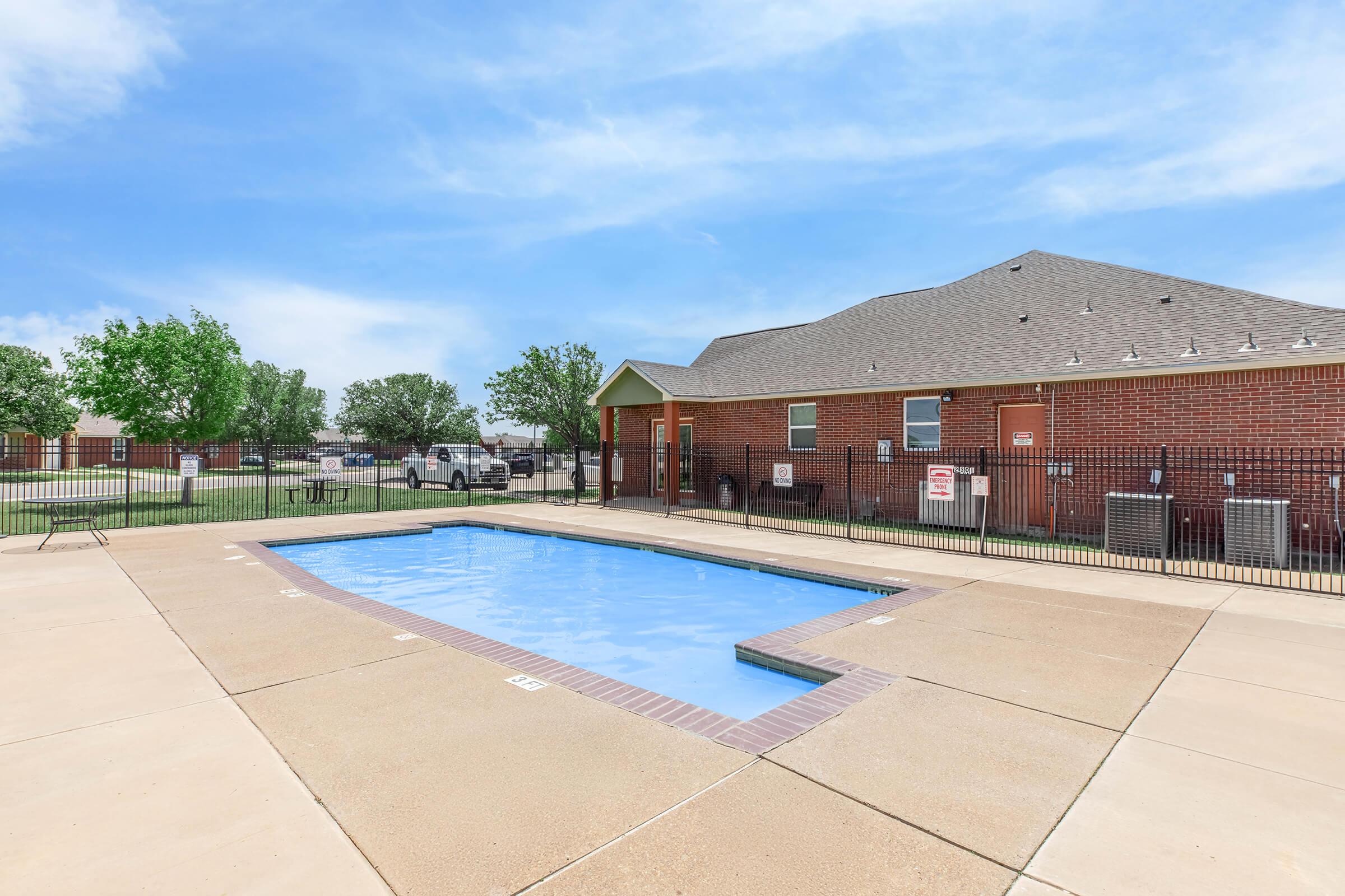A clean, rectangular swimming pool surrounded by concrete. There is a fence around the pool area, and a brick building is visible in the background. Lush green trees can be seen nearby, and the sky is bright with a few clouds.