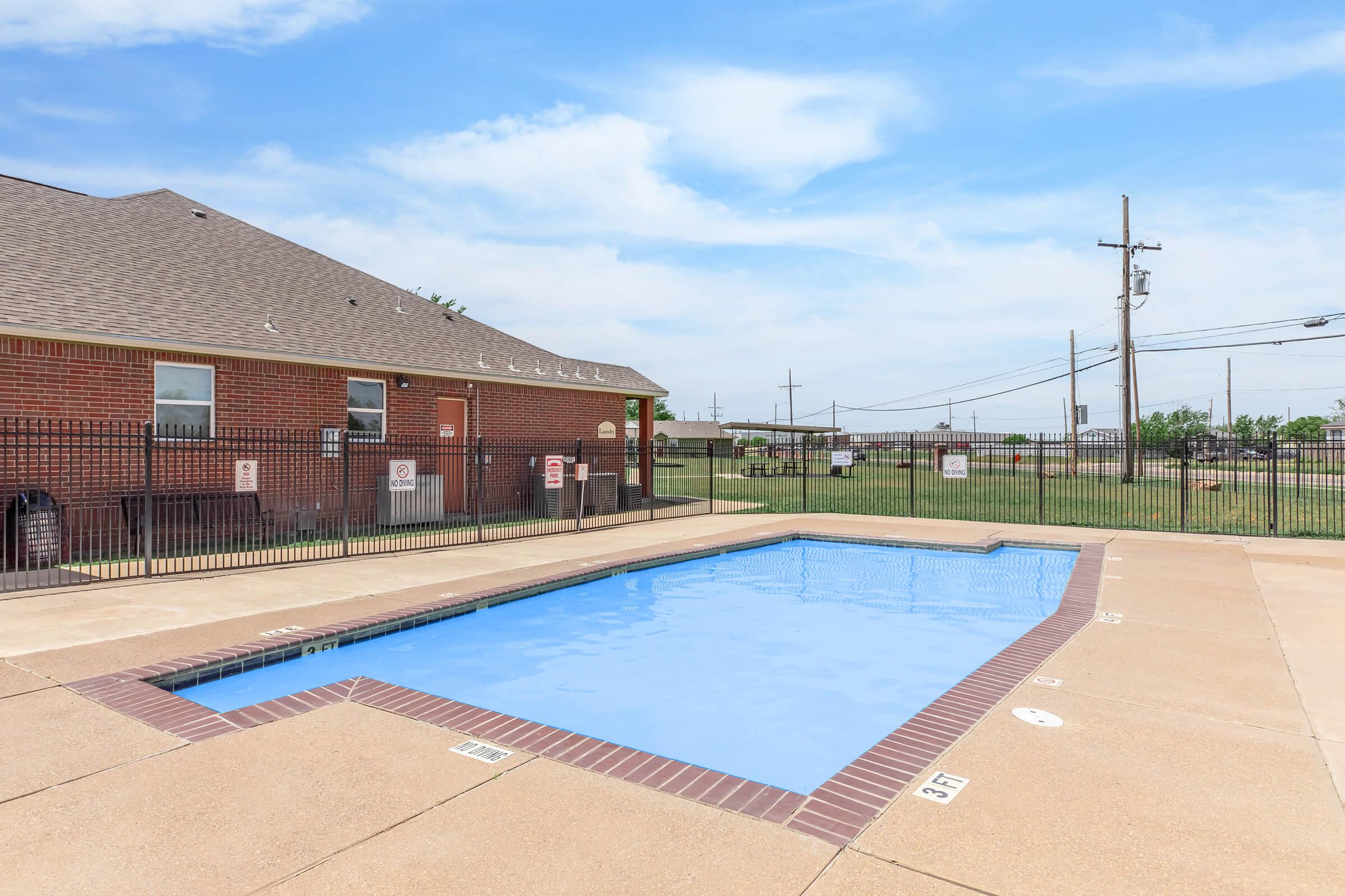 A clear blue swimming pool surrounded by a tan concrete deck. The pool is enclosed by a black metal fence. In the background, there is a brick building with multiple windows and a grassy area. Power lines are visible across the sky, which is partly cloudy.