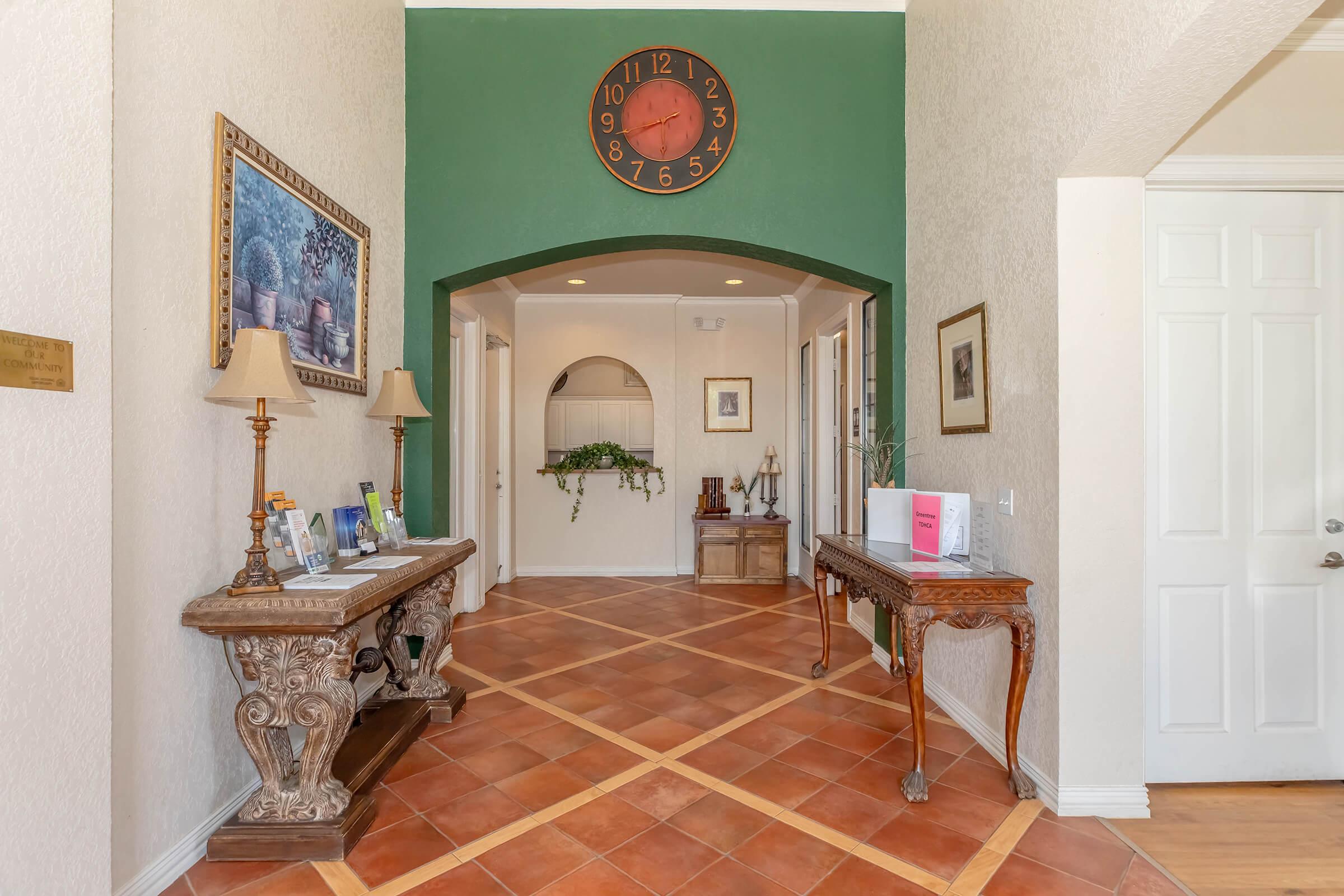 A spacious entryway featuring warm tile flooring, decorative tables with lamps and photo frames, and a large green wall clock. The space is adorned with artwork and plants, creating a welcoming atmosphere. Doorways lead to additional rooms on either side of the hall.