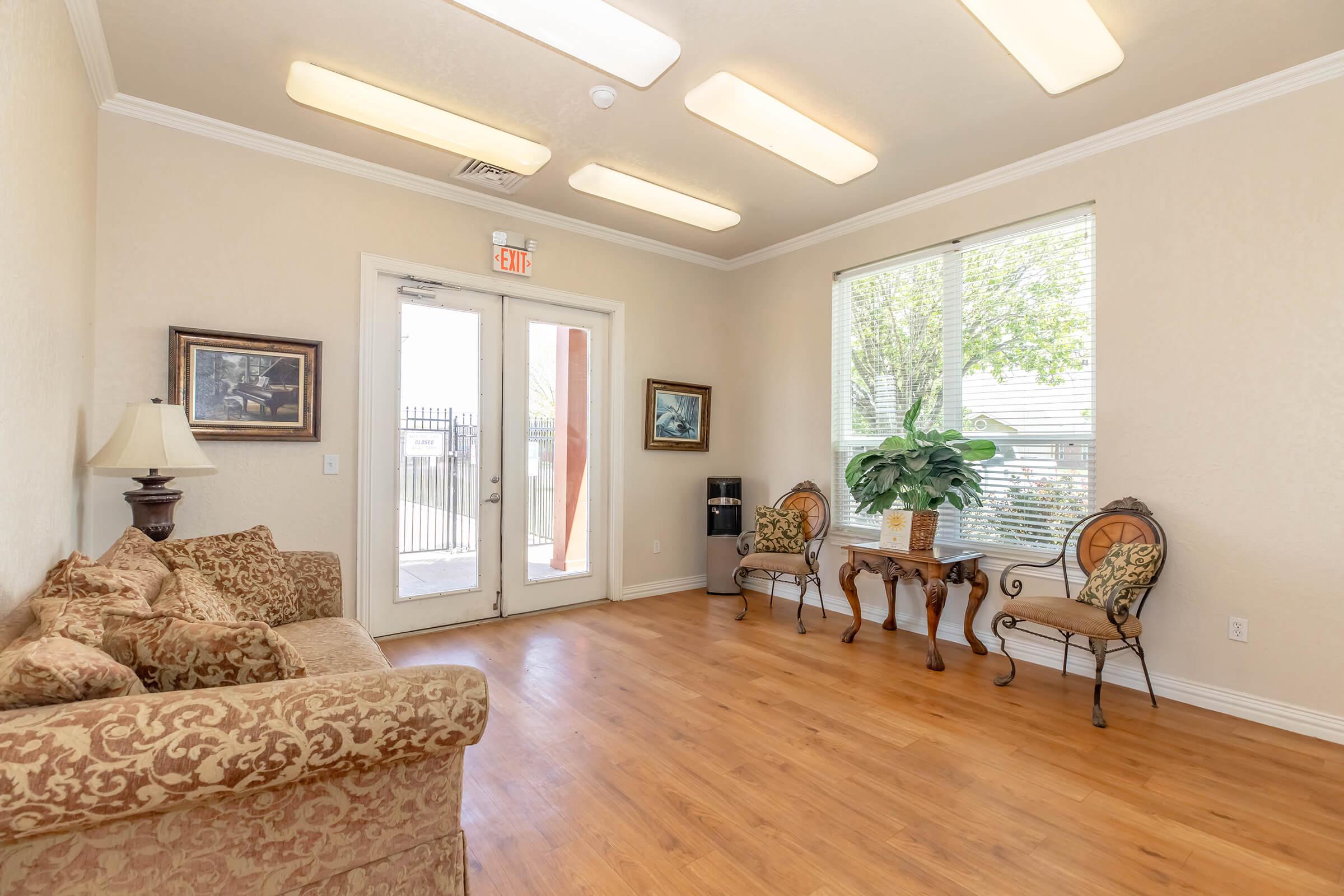 A cozy waiting area with a floral-patterned sofa and two decorative chairs. There's a small wooden table with a plant and informational materials. The room features large windows with natural light, beige walls, and a door leading outside. A lamp adds a warm touch to the space.