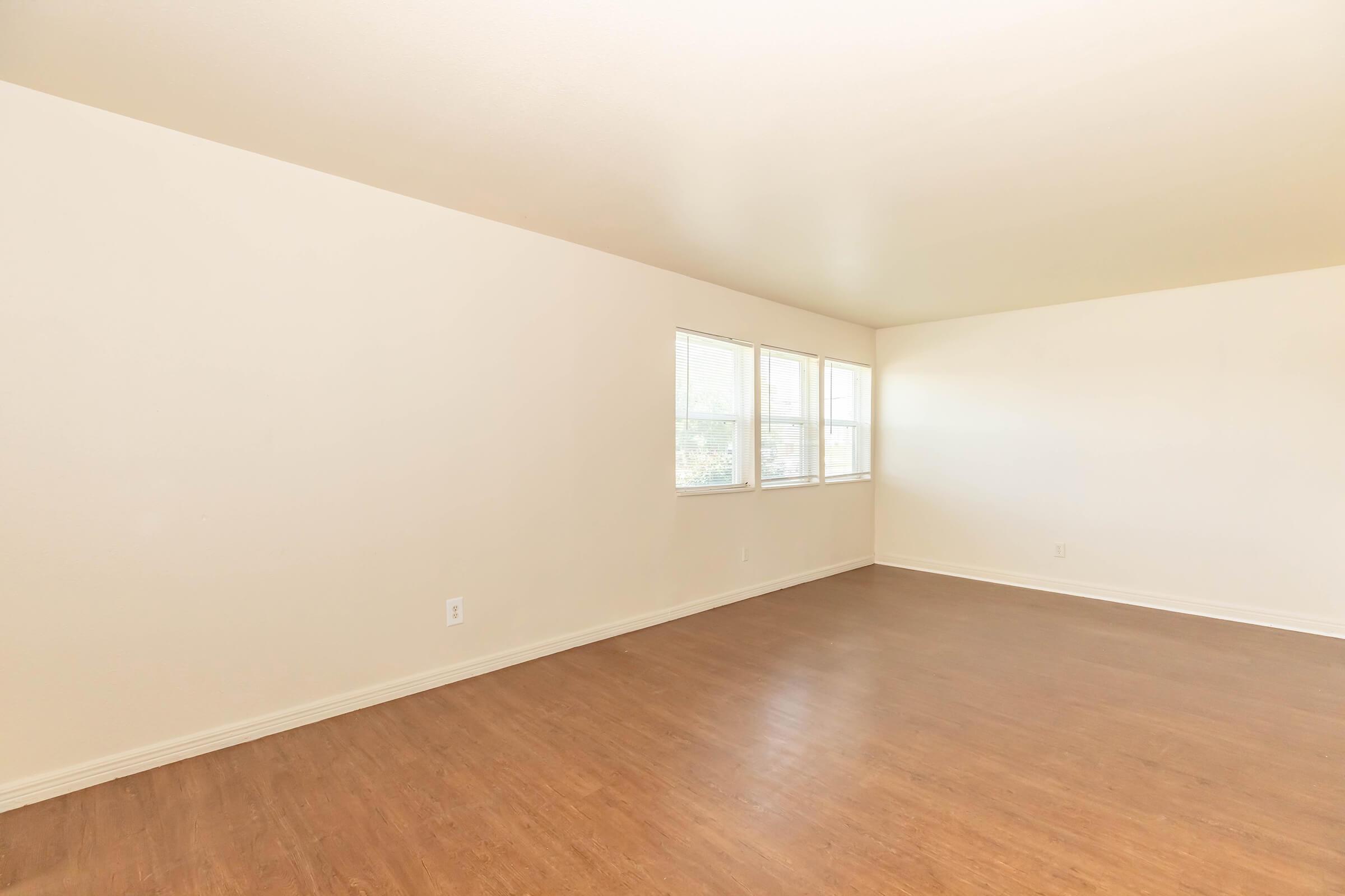 A spacious, empty room with light-colored walls and a wood laminate floor. Three large windows along one wall allow natural light to fill the space. The room has no furniture, creating a bright and open atmosphere, ideal for various layouts or uses.