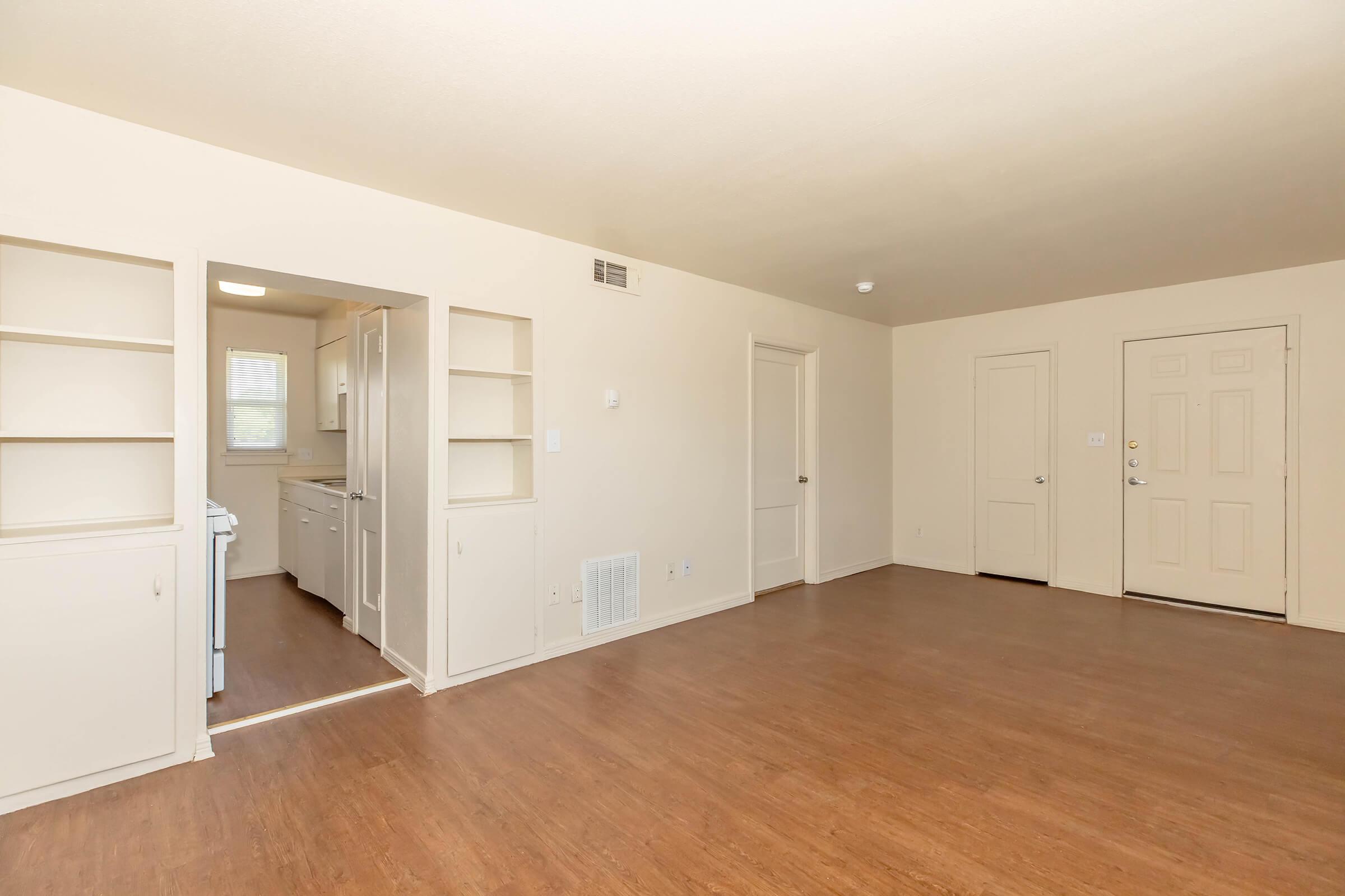 An empty, well-lit living space featuring a wood floor. It includes a kitchen area visible through an open doorway on the left, adjacent built-in shelves, and a single main door on the right. The walls are painted a light color, creating a spacious and inviting atmosphere.