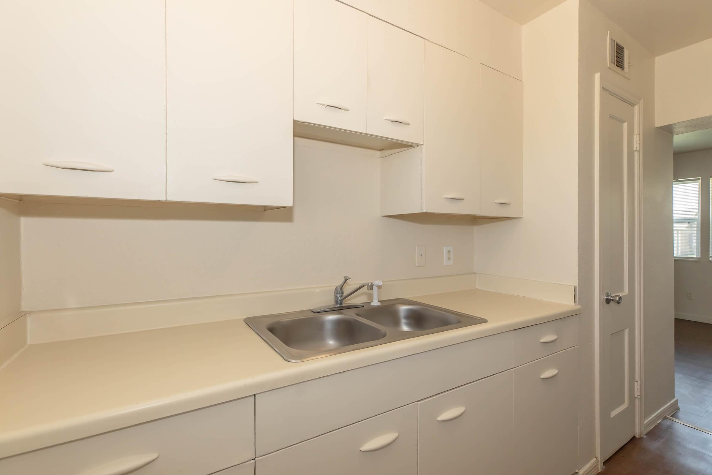 A clean and modern kitchen space featuring beige cabinets overhead, a long countertop, and a stainless steel sink. The walls are painted a light neutral color, and there is a doorway leading to another room in the background. The overall design is simple and functional.