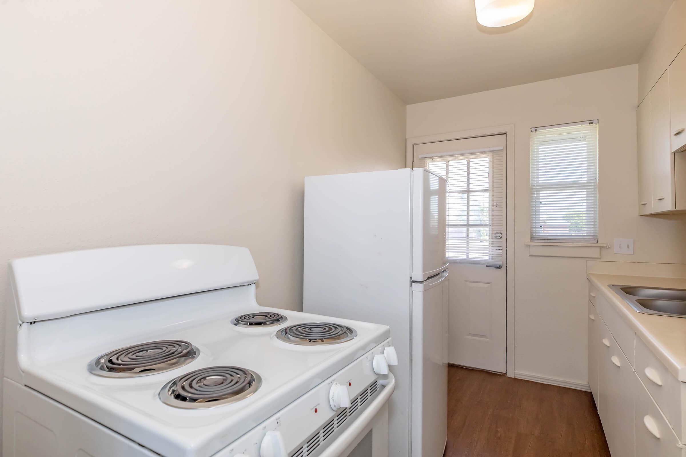 A bright, small kitchen featuring a white stove with four burners, a white refrigerator, and a sink. The walls are light-colored, and there is a door leading outside. Natural light enters through a window with blinds, enhancing the airy feel of the space. Wood laminate flooring adds warmth to the room.